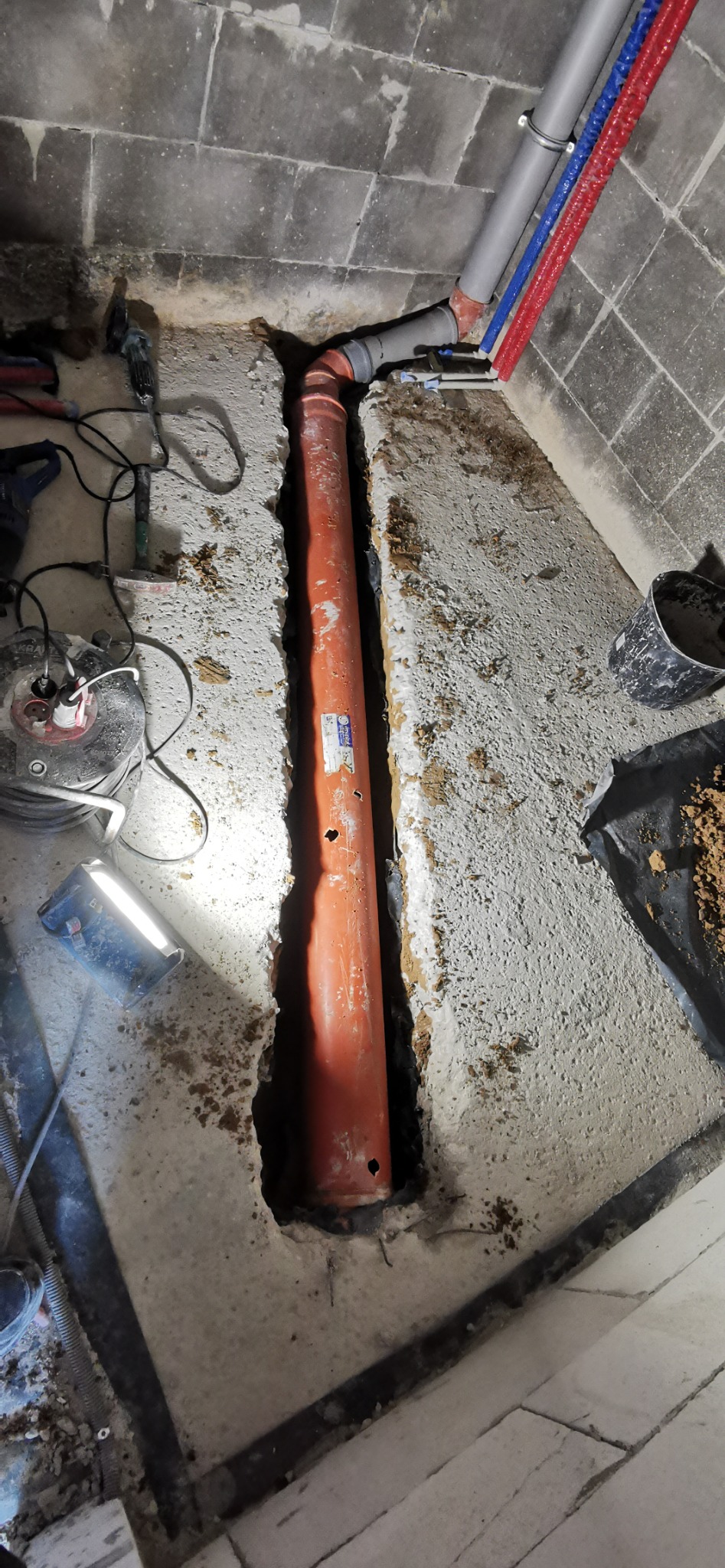 Pomarańczowa rura kanalizacyjna w wykopie w betonowej podłodze, widoczne narzędzia i rury instalacji wodnej. Etap prac instalacyjnych.