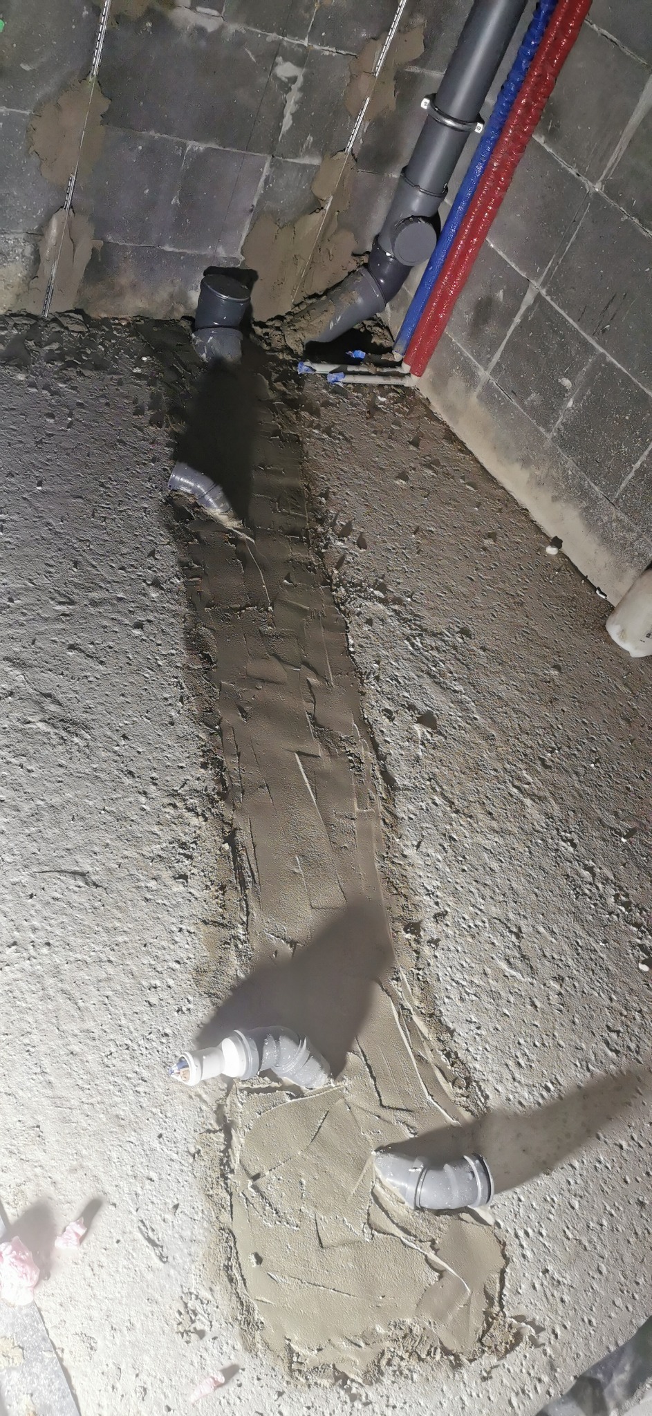 Montaż rur kanalizacyjnych w bruzdach w podłodze, widoczne rury szare, czerwone i niebieskie, podłoże betonowe z zaprawą. Prace hydrauliczne w trakcie realizacji.