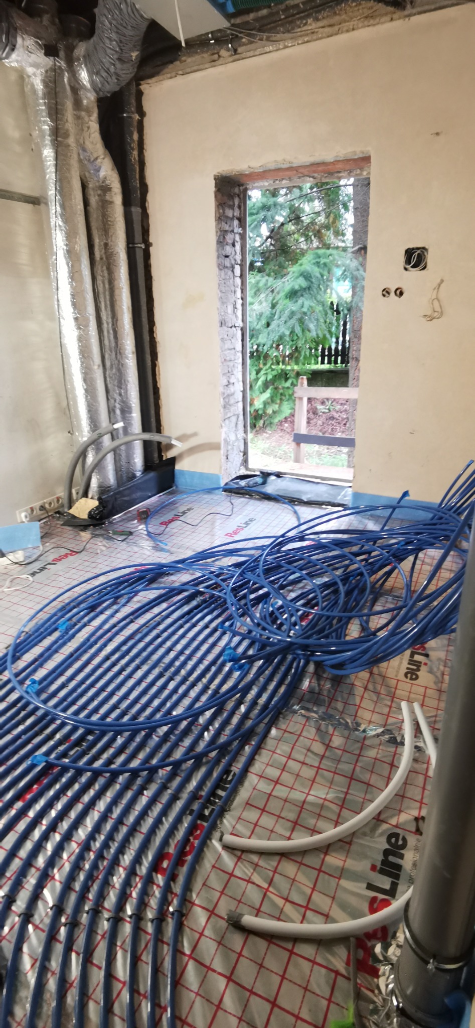 Instalacja ogrzewania podłogowego w trakcie montażu. Widoczne niebieskie rury ułożone na folii izolacyjnej. W tle ściana z otwartym oknem na ogród.