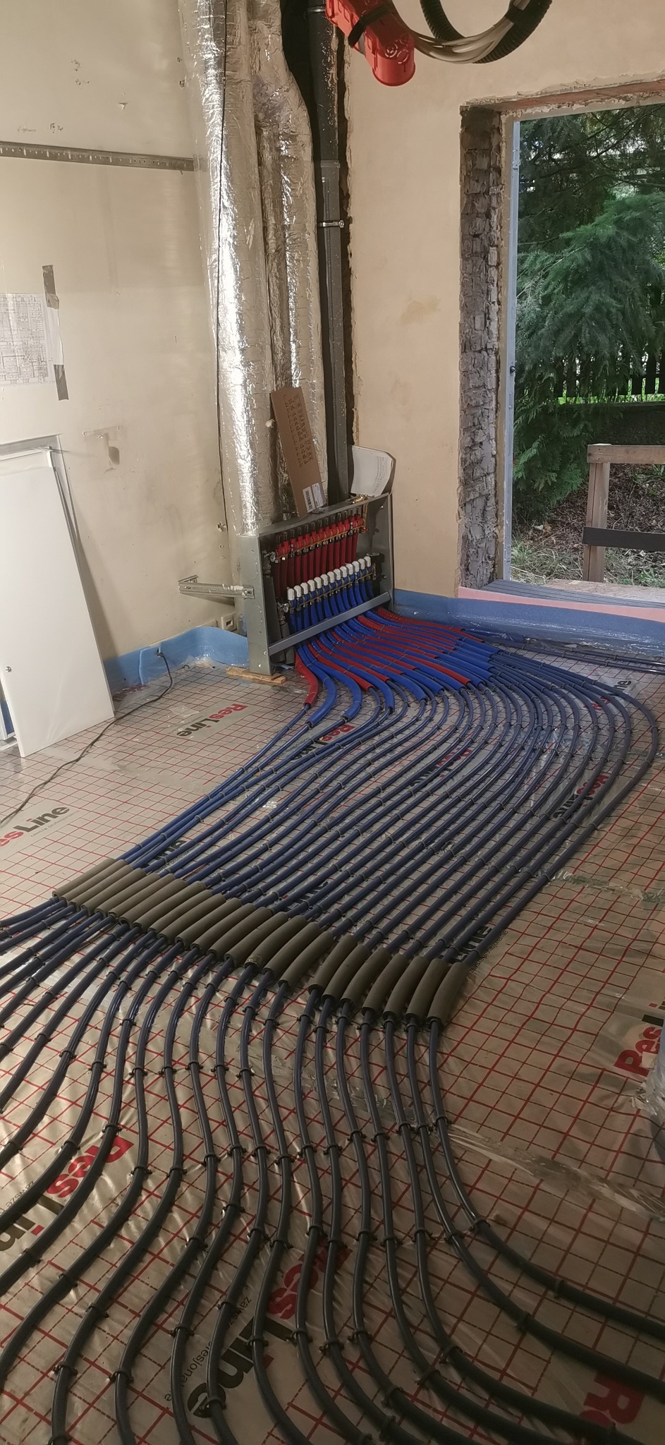Rozłożona instalacja ogrzewania podłogowego z widocznym rozdzielaczem, rurami w kolorze niebieskim i czerwonym, oraz izolacją na części rur. Widok na okno w tle.