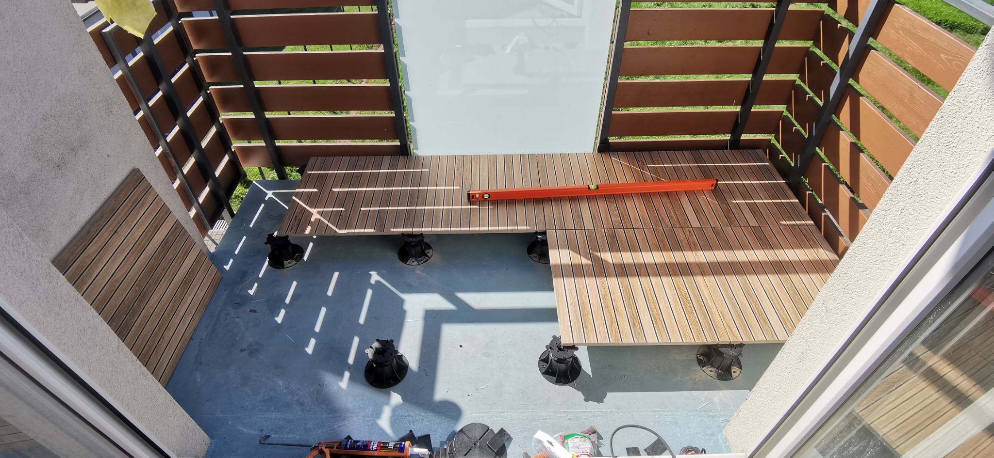 Montaż tarasu drewnianego na regulowanych wspornikach, poziomowany poziomicą, widok z góry na balkon z drewnianą zabudową i fragmentem ogrodzenia.