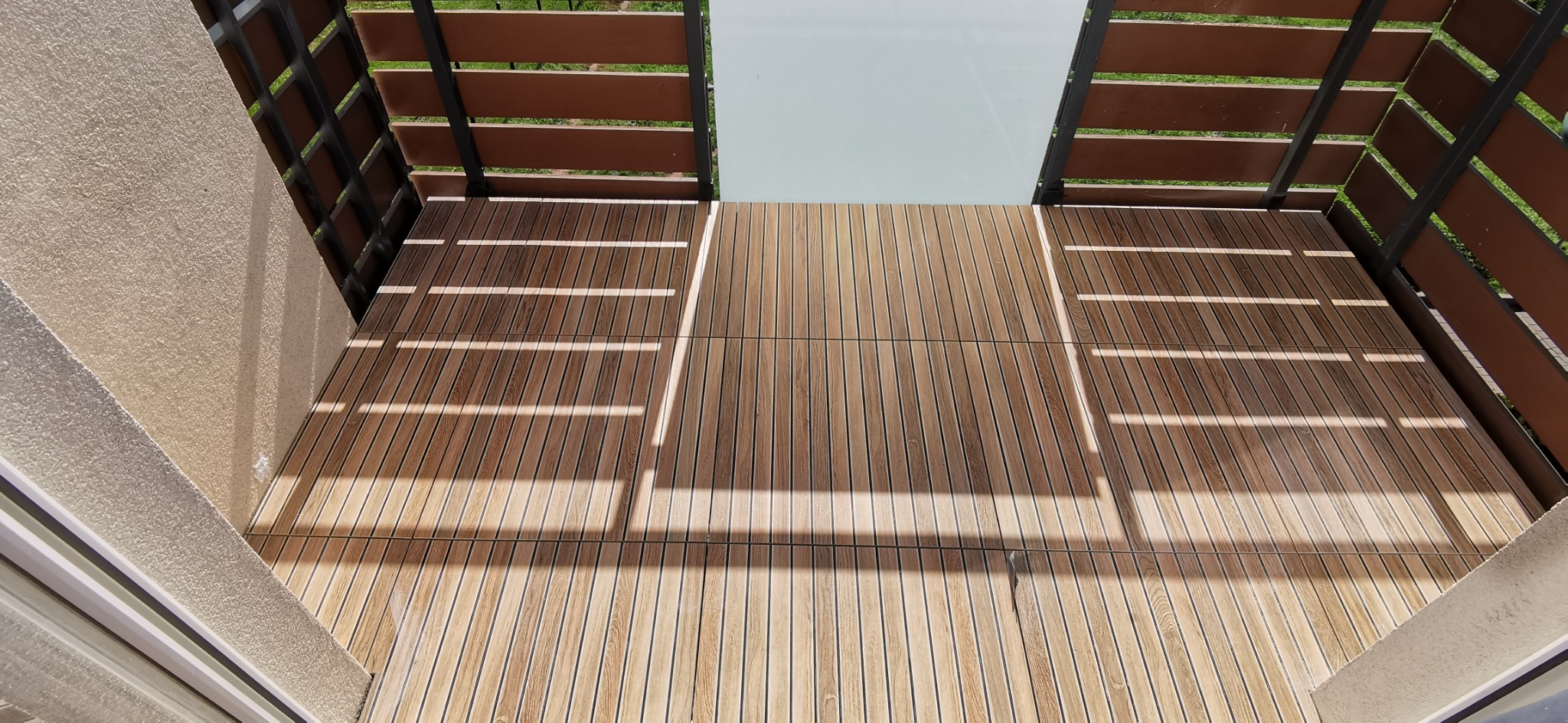 Balkon z drewnianą podłogą z desek kompozytowych, widok z góry, z fragmentami balustrady z brązowych paneli i czarnych metalowych elementów.