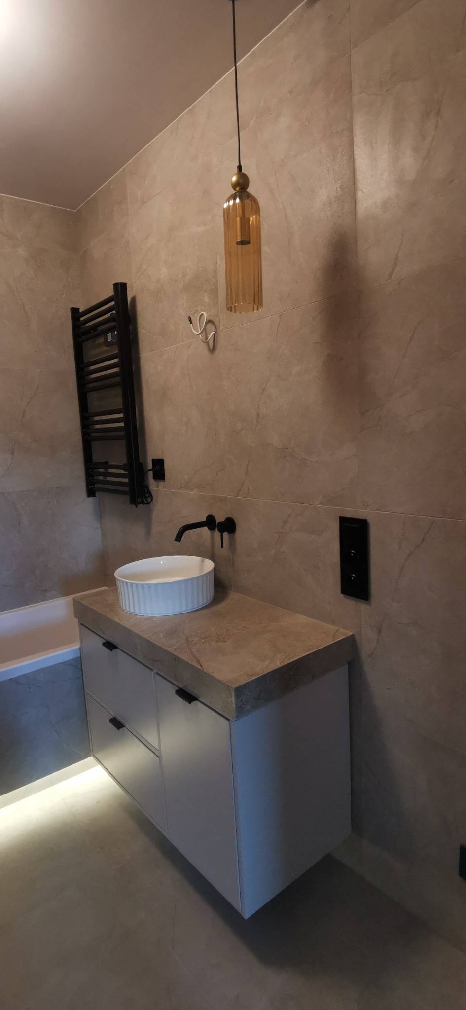 Nowoczesna łazienka z umywalką nablatową, czarną armaturą, grzejnikiem drabinkowym i wiszącą lampą. Stonowana kolorystyka płytek i podświetlenie LED.