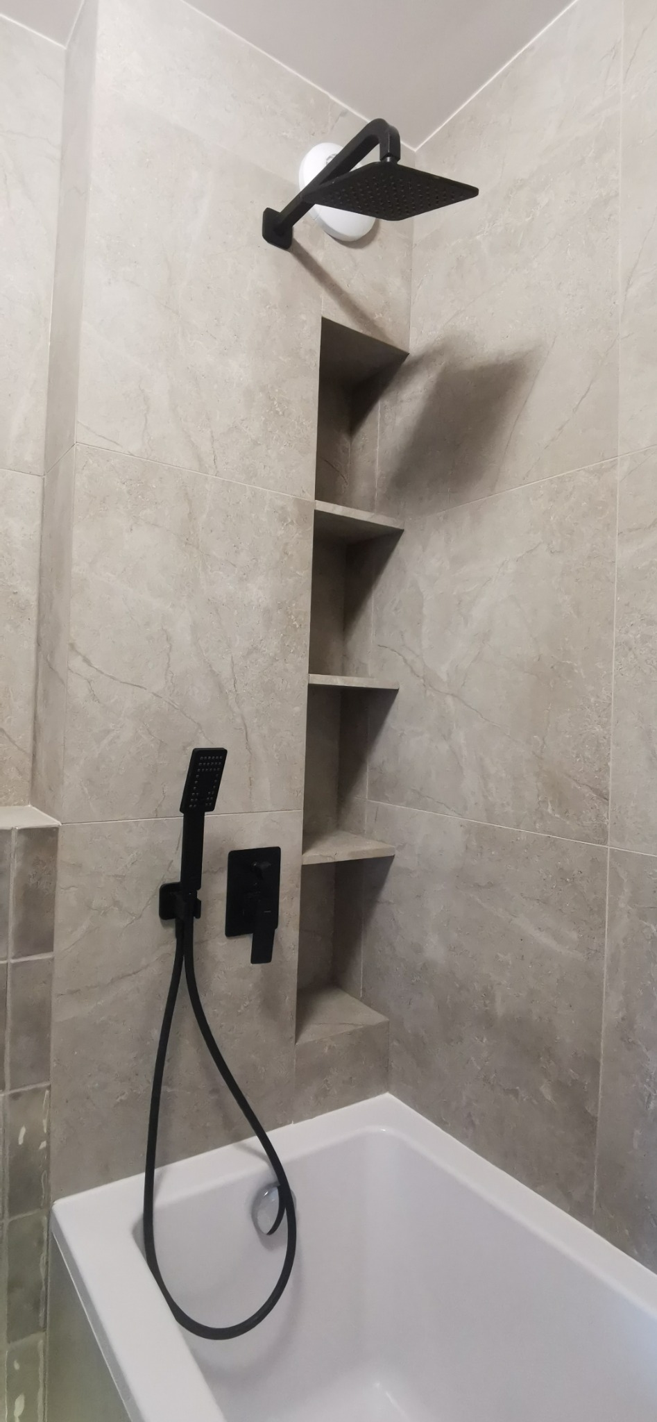 Wykończona łazienka: biała wanna, czarna armatura (deszczownica, słuchawka, bateria) i wnęka z półkami w płytkach imitujących marmur. Nowoczesny design.