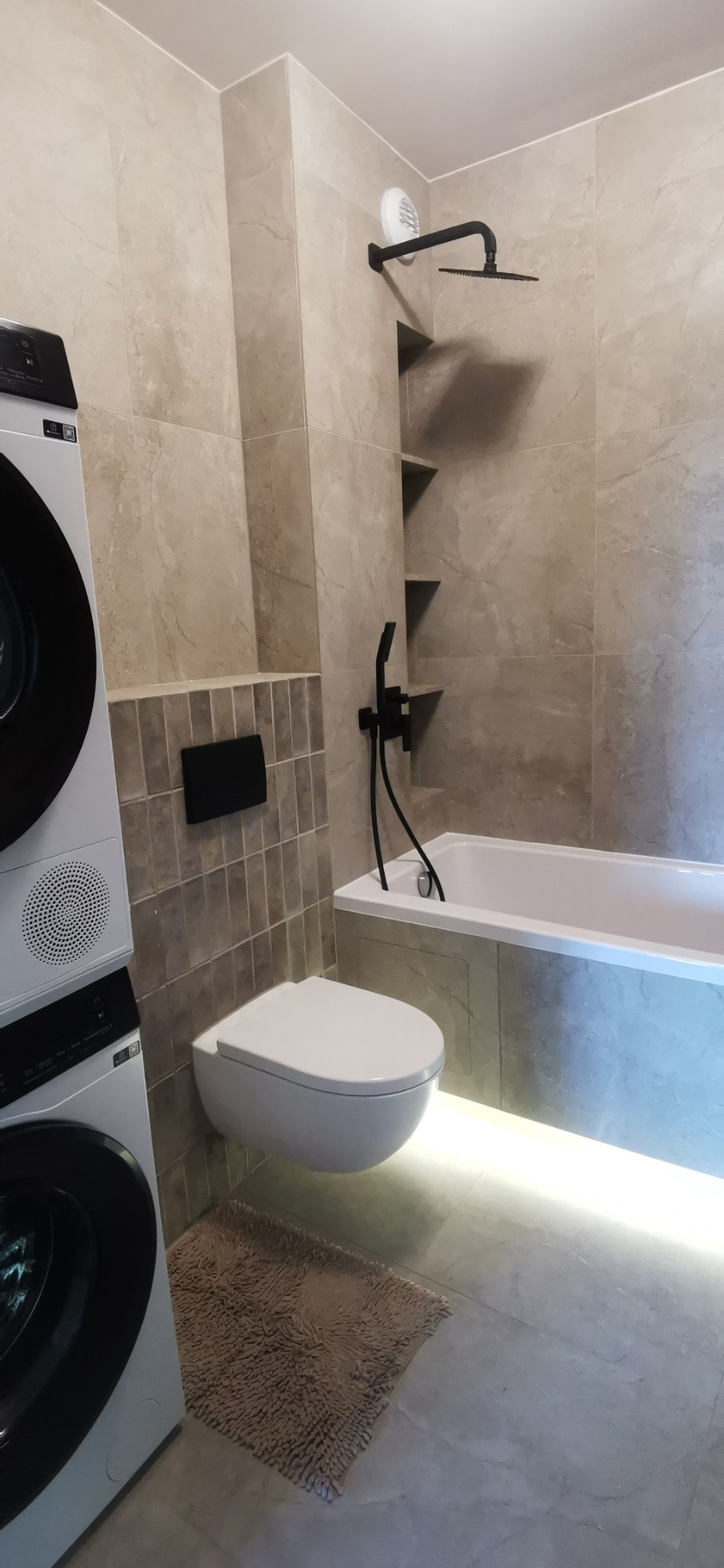 Nowoczesna łazienka z podświetlaną wanną, wiszącą toaletą, czarną armaturą i pralko-suszarką. Płytki imitujące marmur. Minimalistyczny design.
