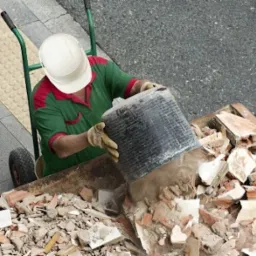 Pracownik w kasku i rękawicach wylewa zawartość czarnego wiadra do kontenera wypełnionego gruzem, widok z góry.