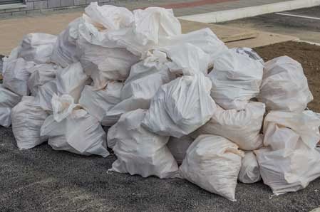 Stos białych, plastikowych worków wypełnionych materiałem budowlanym, leżących na szarym podłożu z drobnymi kamykami, z fragmentem krawężnika w tle.