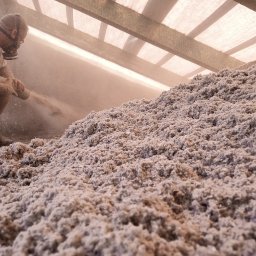 TOMASZ KOPERTOWSKI TOMKOP - Pracownik w masce ochronnej aplikuje sypką izolację na poddasze za pomocą węża, widoczna gruba warstwa materiału izolacyjnego i konstrukcja dachu.