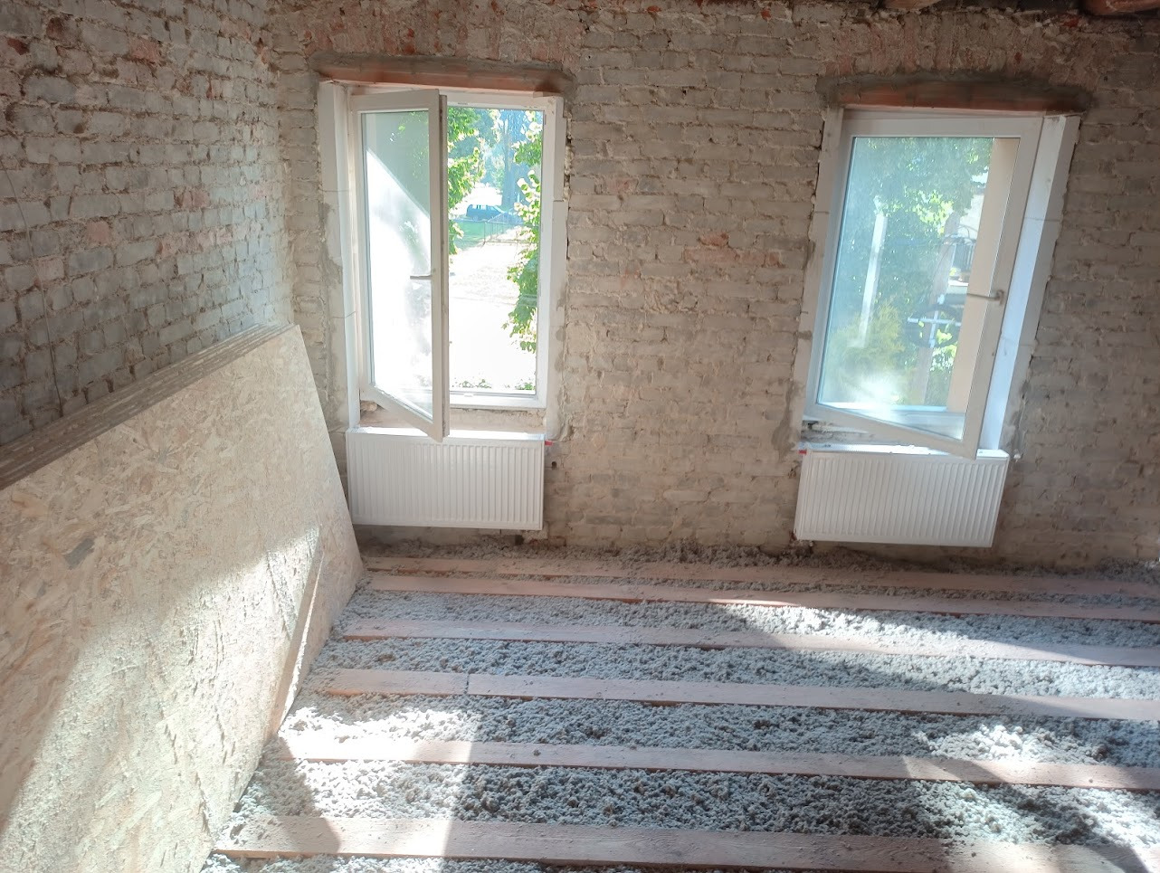 Poddasze w trakcie prac izolacyjnych: widoczne deski podłogowe, biała wełna mineralna między nimi, stos płyt OSB przy ścianie z cegły oraz dwa białe okna połaciowe z grzejnikami. Ściany w stanie...