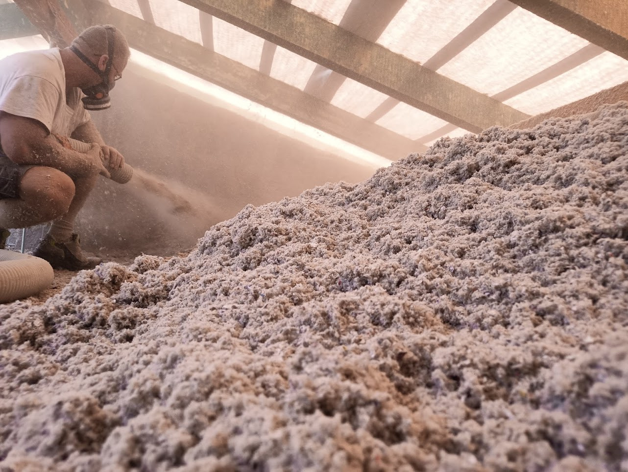 Pracownik w masce ochronnej aplikuje sypką izolację na poddasze za pomocą węża, widoczna gruba warstwa materiału izolacyjnego i konstrukcja dachu.
