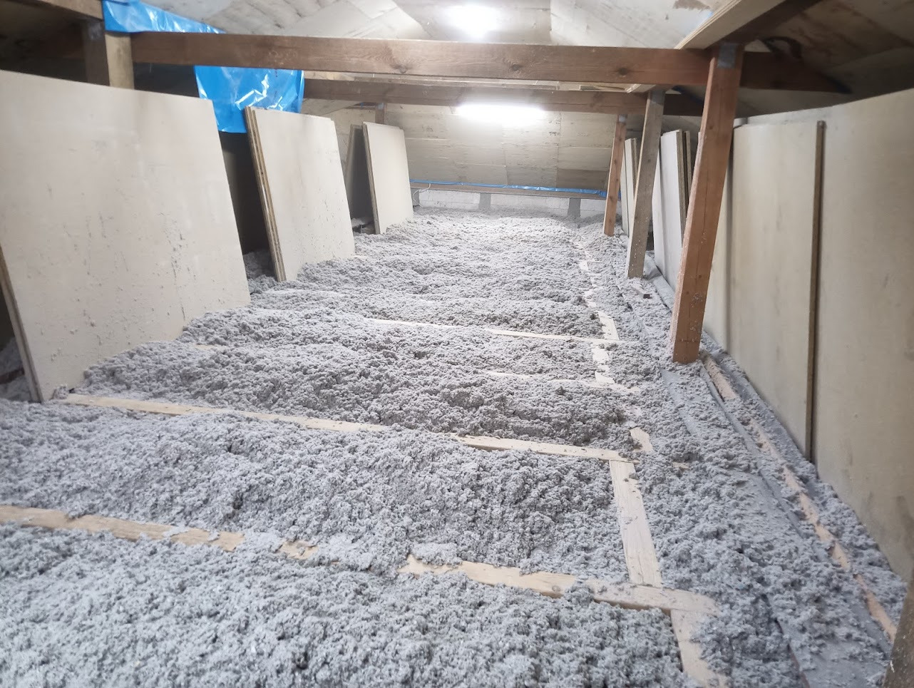 Poddasze z widoczną drewnianą konstrukcją pokryte warstwą szarej izolacji z włókien, częściowo zakryte płytami.