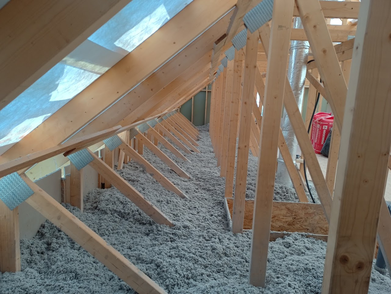 Ocieplenie poddasza wełną celulozową, widoczne drewniane krokwie i elementy konstrukcyjne, folia paroizolacyjna na górze, czerwone opakowanie wełny Rockwool w tle.
