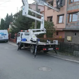 Biały podnośnik koszowy na ulicy przed budynkiem, z pracownikiem w koszu na wysokości okna, widoczny samochód dostawczy Eko Okna w tle.