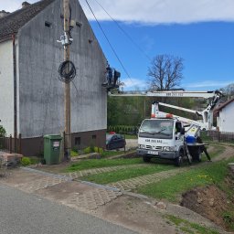 HUSARZ MARIUSZ HUSAK - Praca na elewacji domu z użyciem podnośnika koszowego. Pracownik w koszu na wysokości, biały samochód z podnośnikiem na trawniku. Widok na szarą elewację i słup elektryczny.