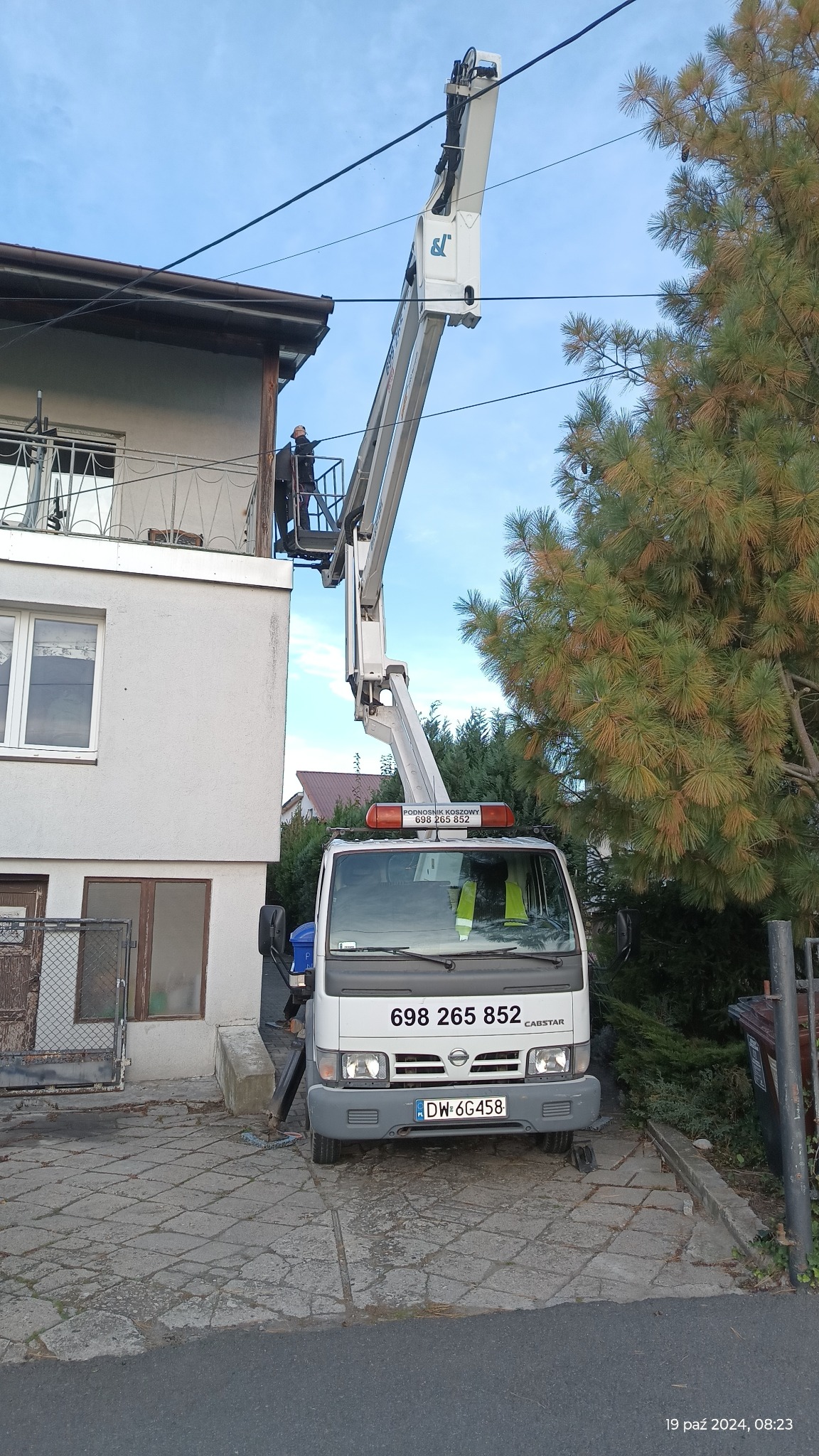 Biały podnośnik koszowy z operatorem w koszu, ustawiony przy dwupiętrowym budynku z balkonem, widoczny numer telefonu na kabinie i tablica rejestracyjna pojazdu.