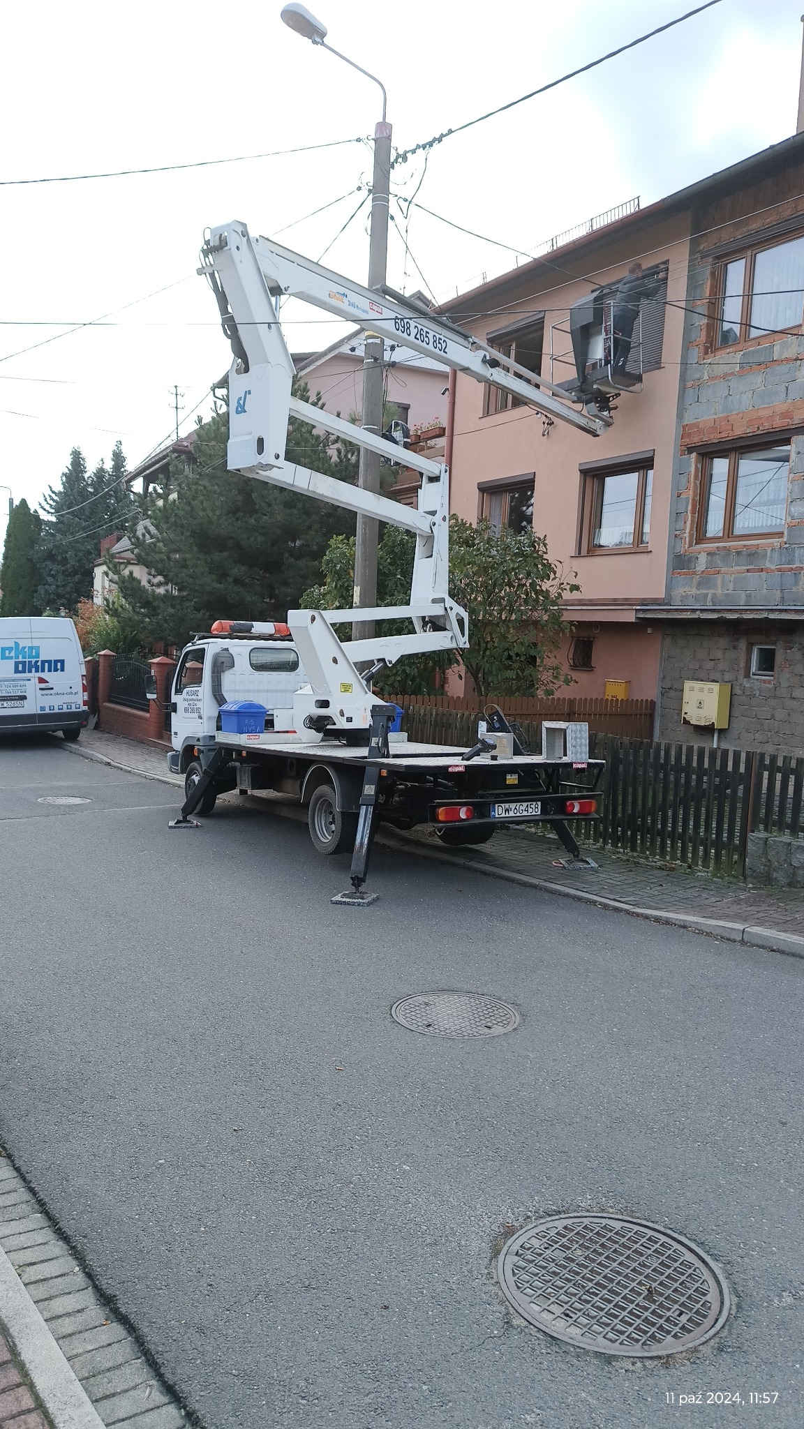 Biały podnośnik koszowy na ulicy przed budynkiem, z pracownikiem w koszu na wysokości okna, widoczny samochód dostawczy Eko Okna w tle.