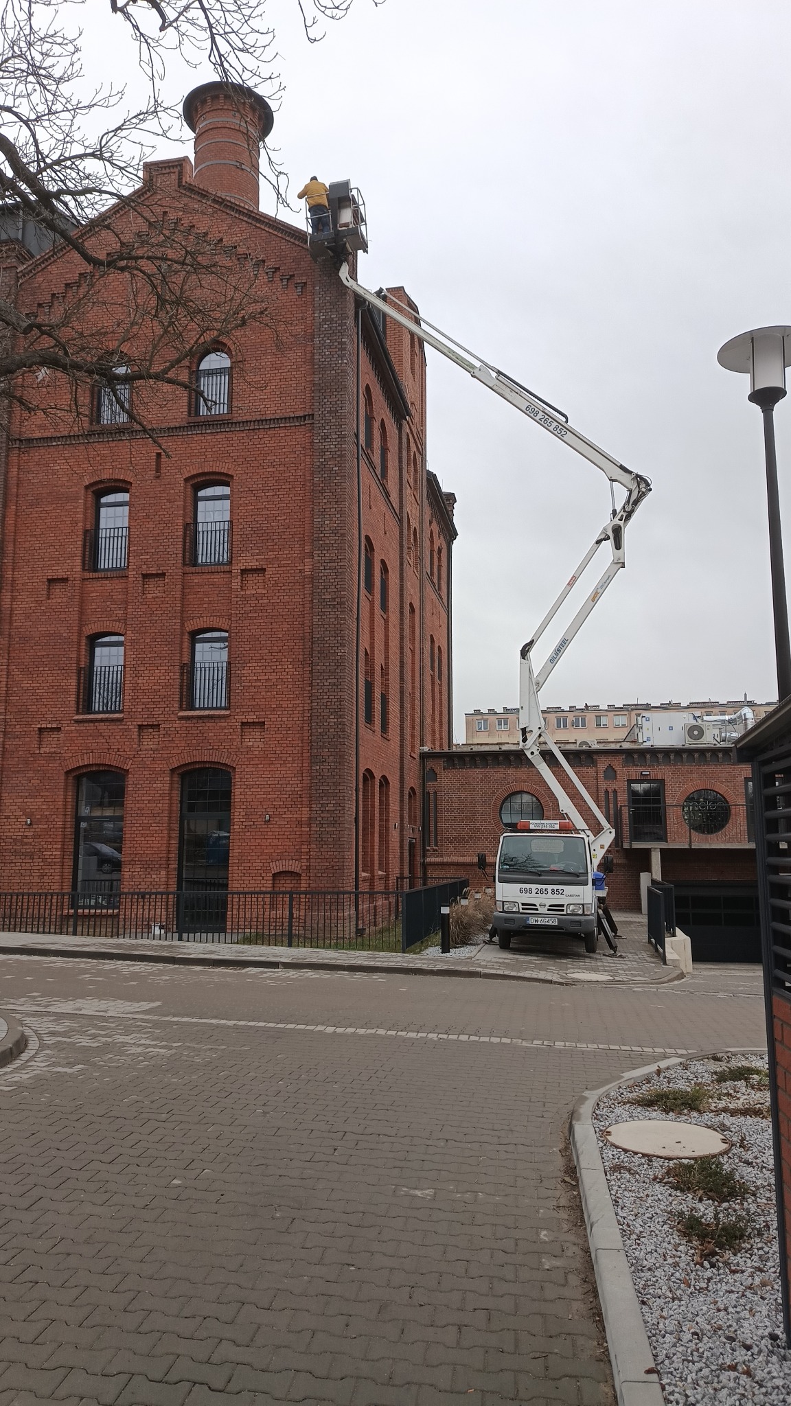 Pracownik w żółtej kurtce w podnośniku koszowym przy ceglanym budynku, prawdopodobnie kontrola lub naprawa komina, na dole widoczny biały samochód z numerem telefonu.