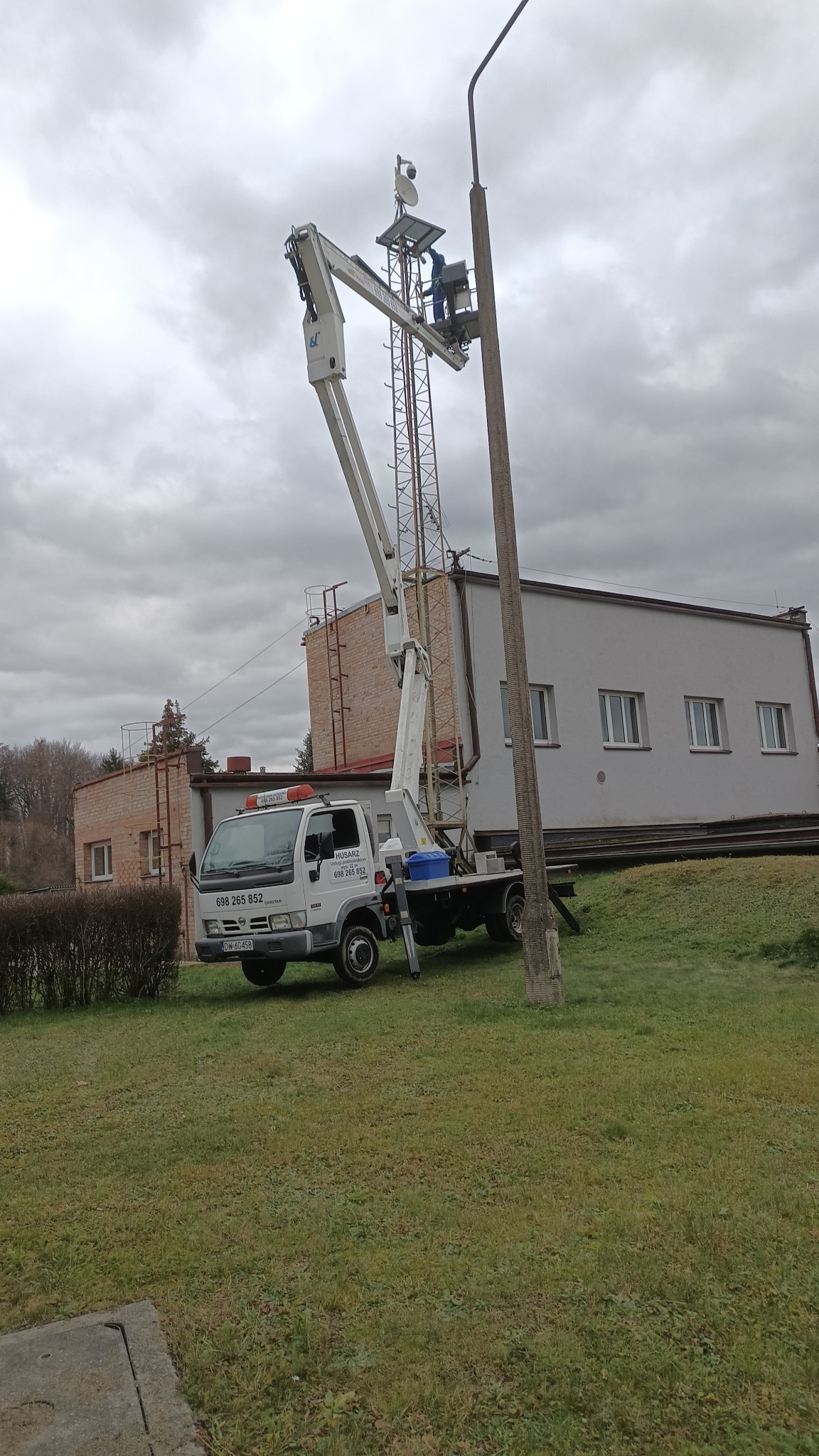 Pracownik w koszu podnośnika teleskopowego, wykonujący prace na wysokości przy maszcie telekomunikacyjnym, na tle pochmurnego nieba i budynku.