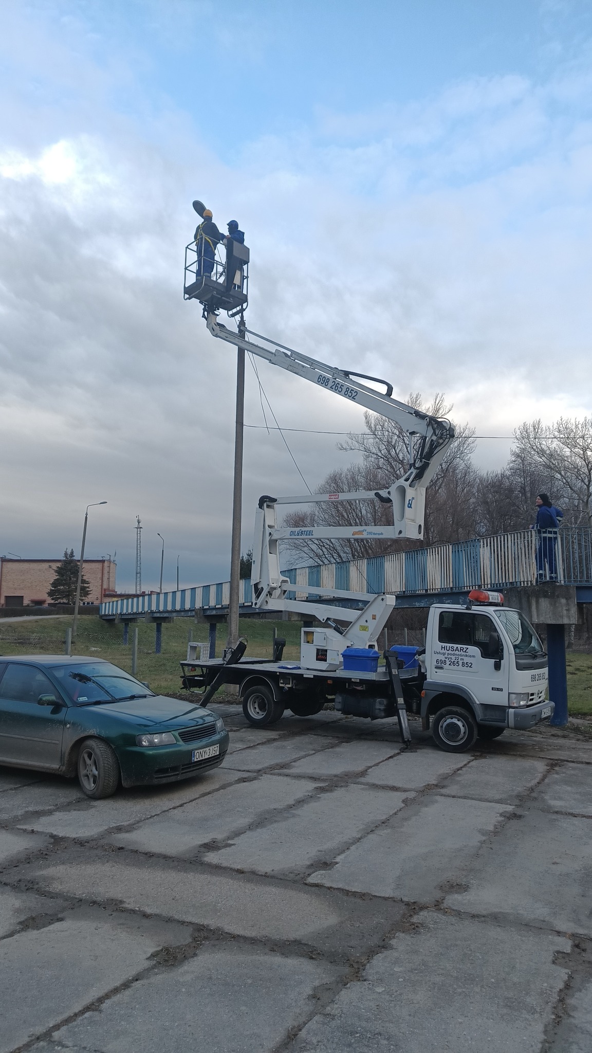 Pracownicy w koszu podnośnika koszowego, na tle pochmurnego nieba, przy słupie oświetleniowym, z widoczną platformą roboczą i samochodem firmy Husarz z numerem telefonu.