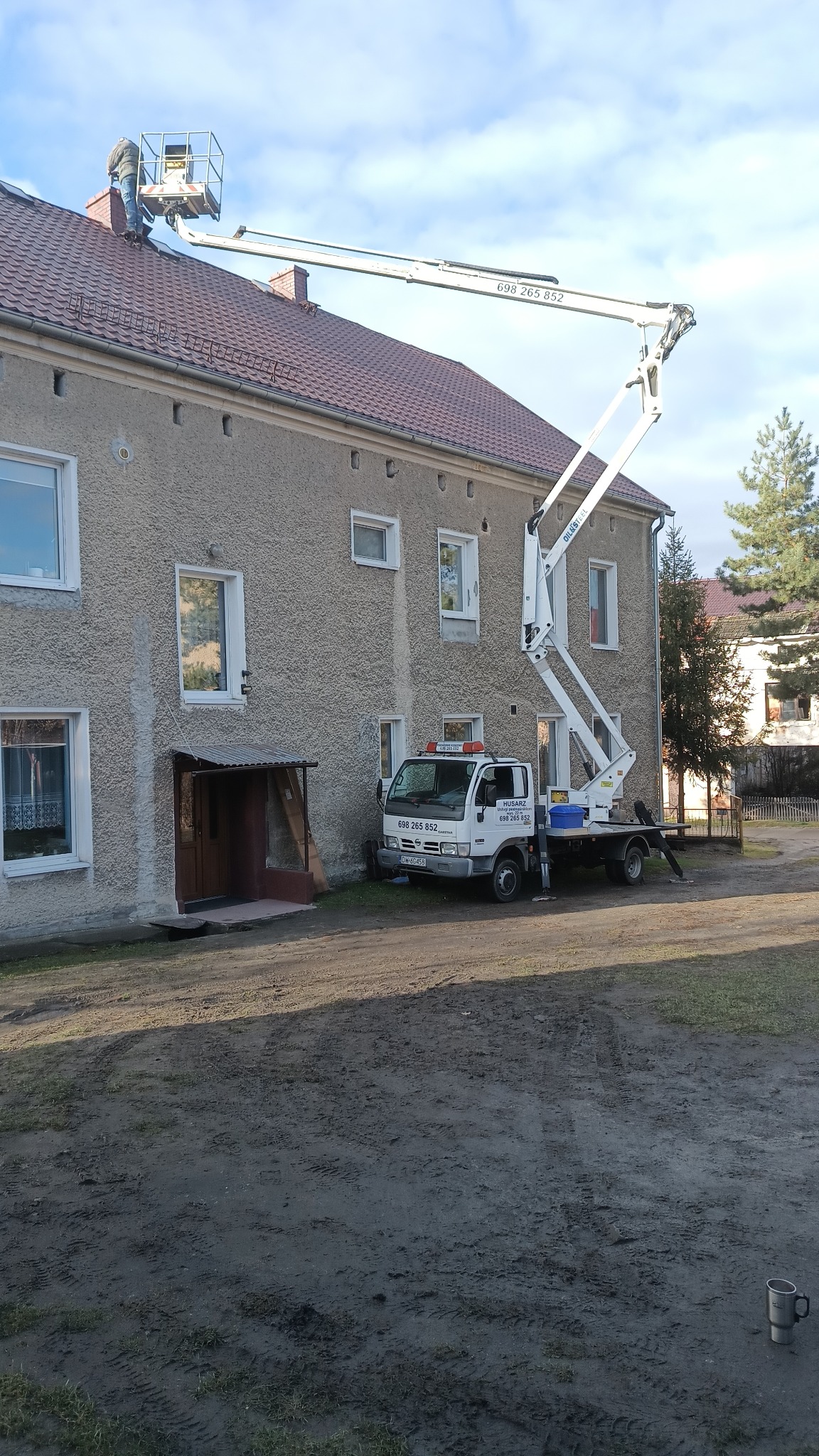 Pracownik na podnośniku koszowym naprawia dach budynku mieszkalnego, widoczny samochód z podnośnikiem oraz fragment podwórka z kubkiem.
