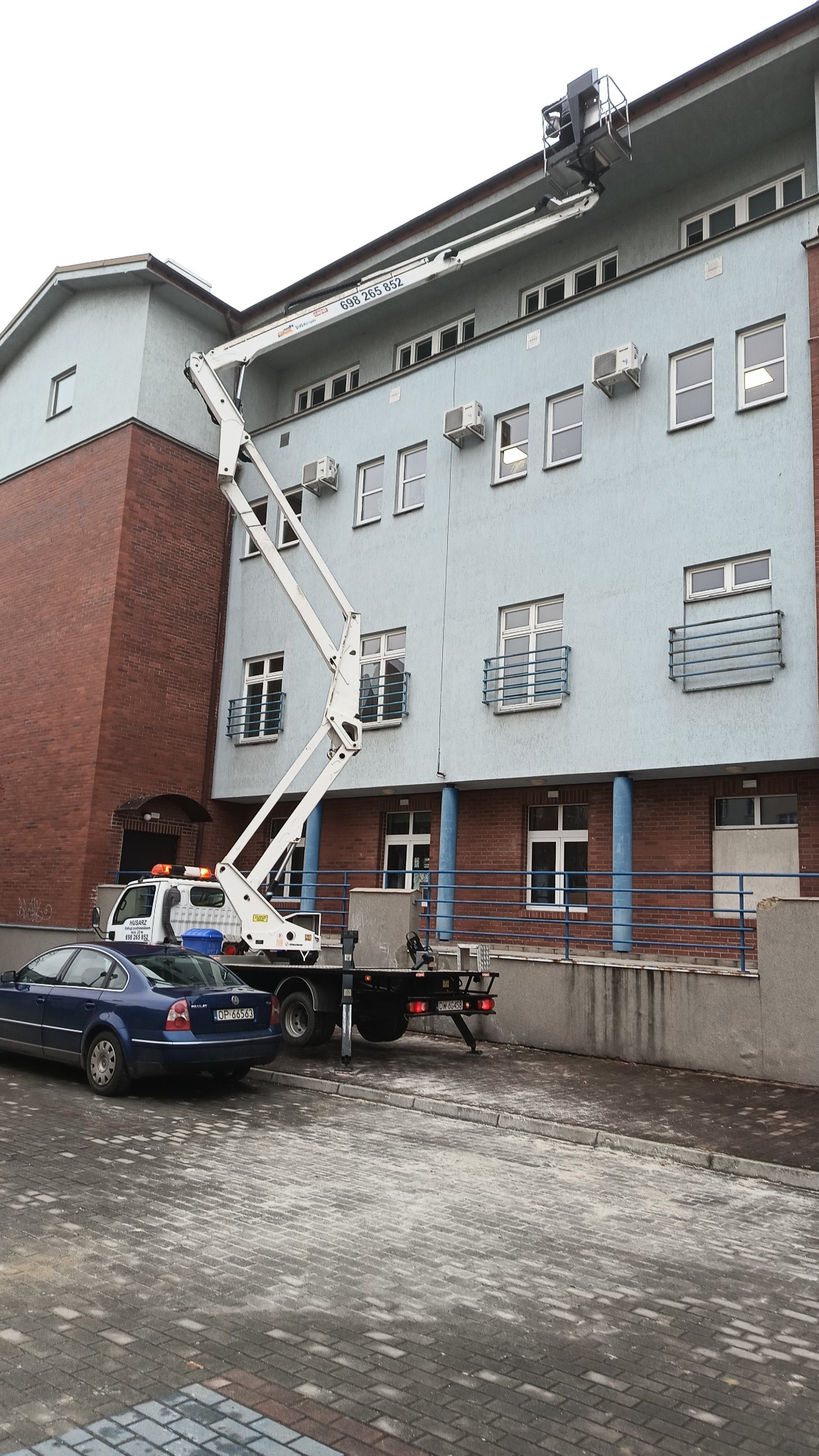 Podnośnik koszowy ustawiony przy budynku z klimatyzatorami, widoczny samochód dostawczy i osobowy na brukowanym placu.