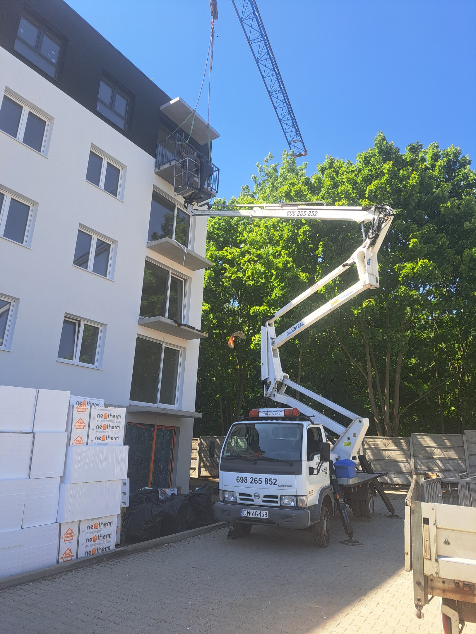 Montaż balkonu w bloku przy użyciu podnośnika koszowego. Widoczne materiały izolacyjne i fragment żurawia w tle.