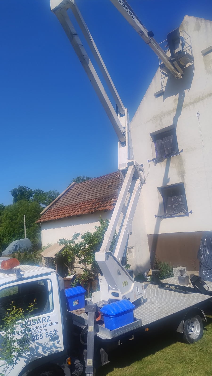 Podnośnik koszowy na tle białej ściany budynku z zabezpieczonymi oknami, w słoneczny dzień. Widoczny fragment dachu sąsiedniego budynku.