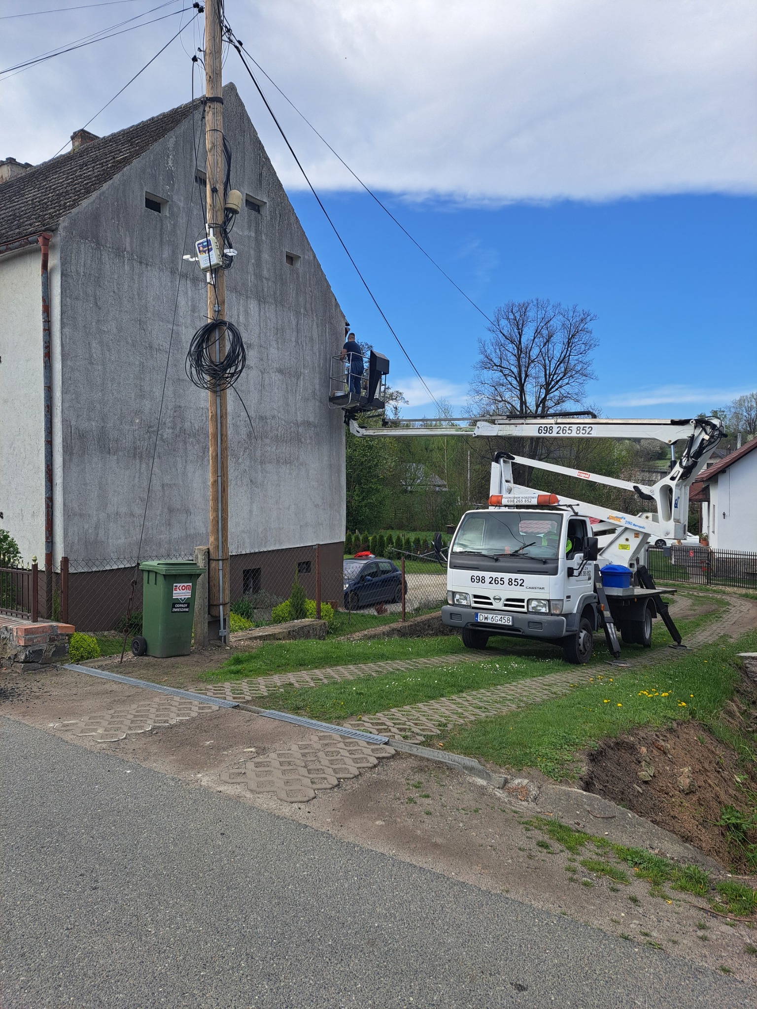 Praca na elewacji domu z użyciem podnośnika koszowego. Pracownik w koszu na wysokości, biały samochód z podnośnikiem na trawniku. Widok na szarą elewację i słup elektryczny.