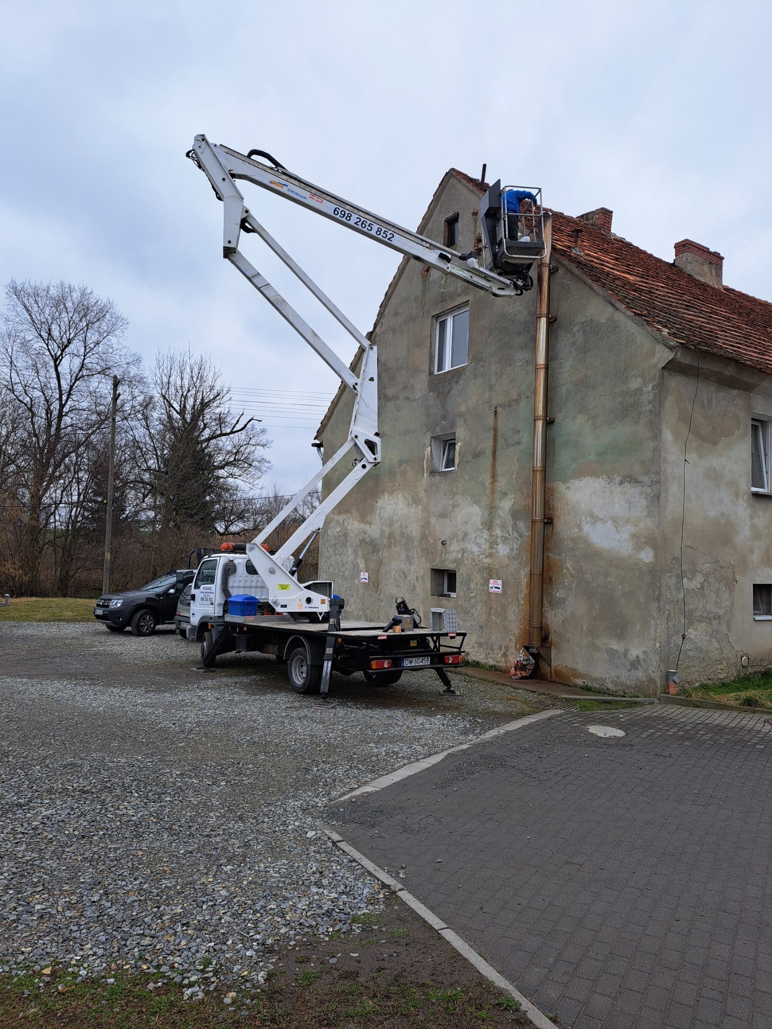 Podnośnik koszowy przy dwukondygnacyjnym budynku z czerwonym dachem. Pracownik w koszu przy ścianie. Szary, pochmurny dzień. Widoczny samochód i drzewa.