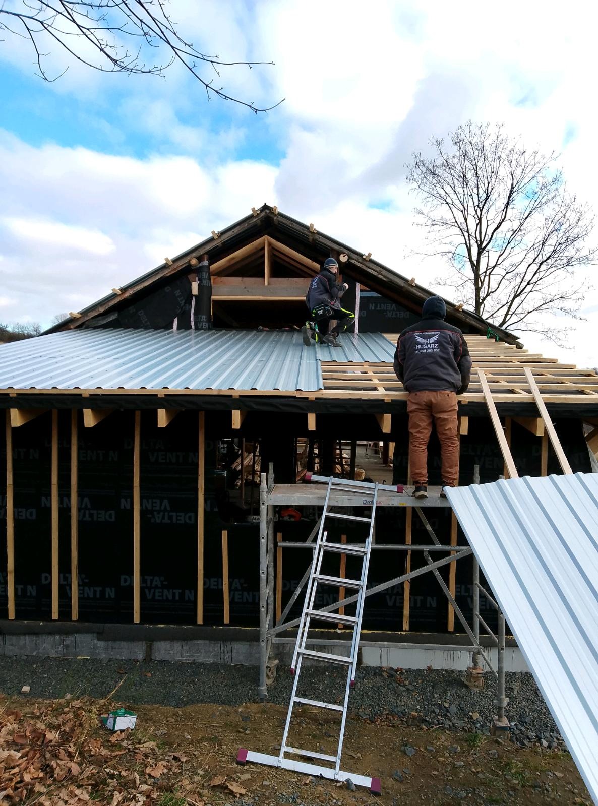 Dwóch dekarzy montuje blachodachówkę na dachu domu jednorodzinnego. Widoczne rusztowania, drabina i elementy konstrukcji dachu. Praca w toku, pochmurne niebo w tle.