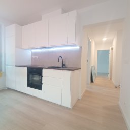 FERMONT USŁUGI BUDOWLANE Wojciech Nowak - Minimalistyczna, biała zabudowa kuchenna z czarną armaturą i piekarnikiem w nowym mieszkaniu, widok na jasny parkiet i korytarz w tle.