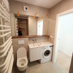 FERMONT USŁUGI BUDOWLANE Wojciech Nowak - Wykończona łazienka z toaletą, umywalką w zabudowie, pralką oraz grzejnikiem drabinkowym, widoczne taśmy malarskie na lustrze.