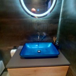 FERMONT USŁUGI BUDOWLANE Wojciech Nowak - Niebieska, szklana umywalka nablatowa w nowoczesnej łazience w Myślenicach, z podświetlanym lustrem i szarą ścianą o fakturze betonu.