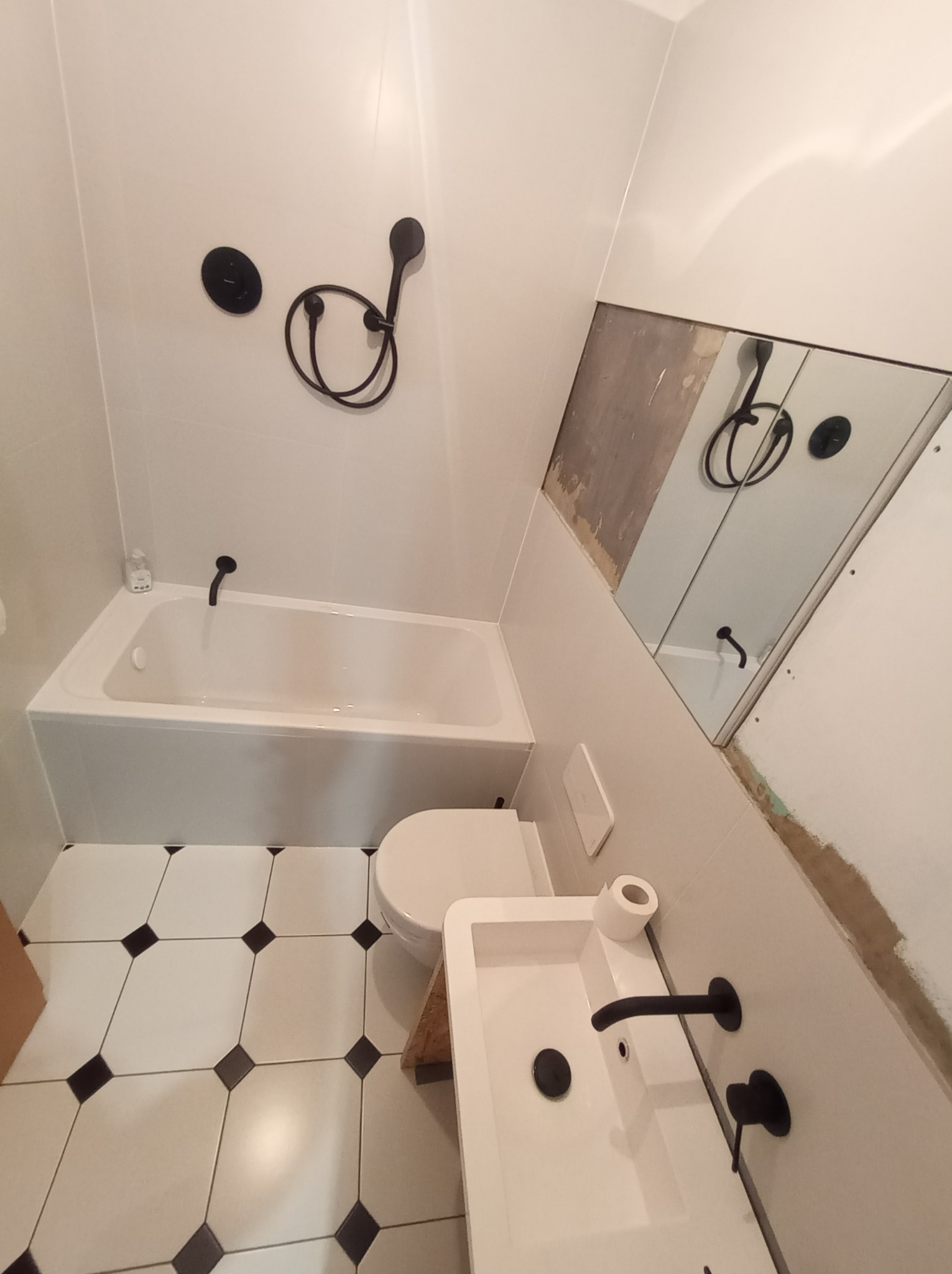 Ukończona łazienka z białą armaturą i czarnymi detalami, widoczne płytki podłogowe w biało-czarną szachownicę, bateria umywalkowa i prysznicowa w kolorze czarnym, częściowo odsłonięta ściana...