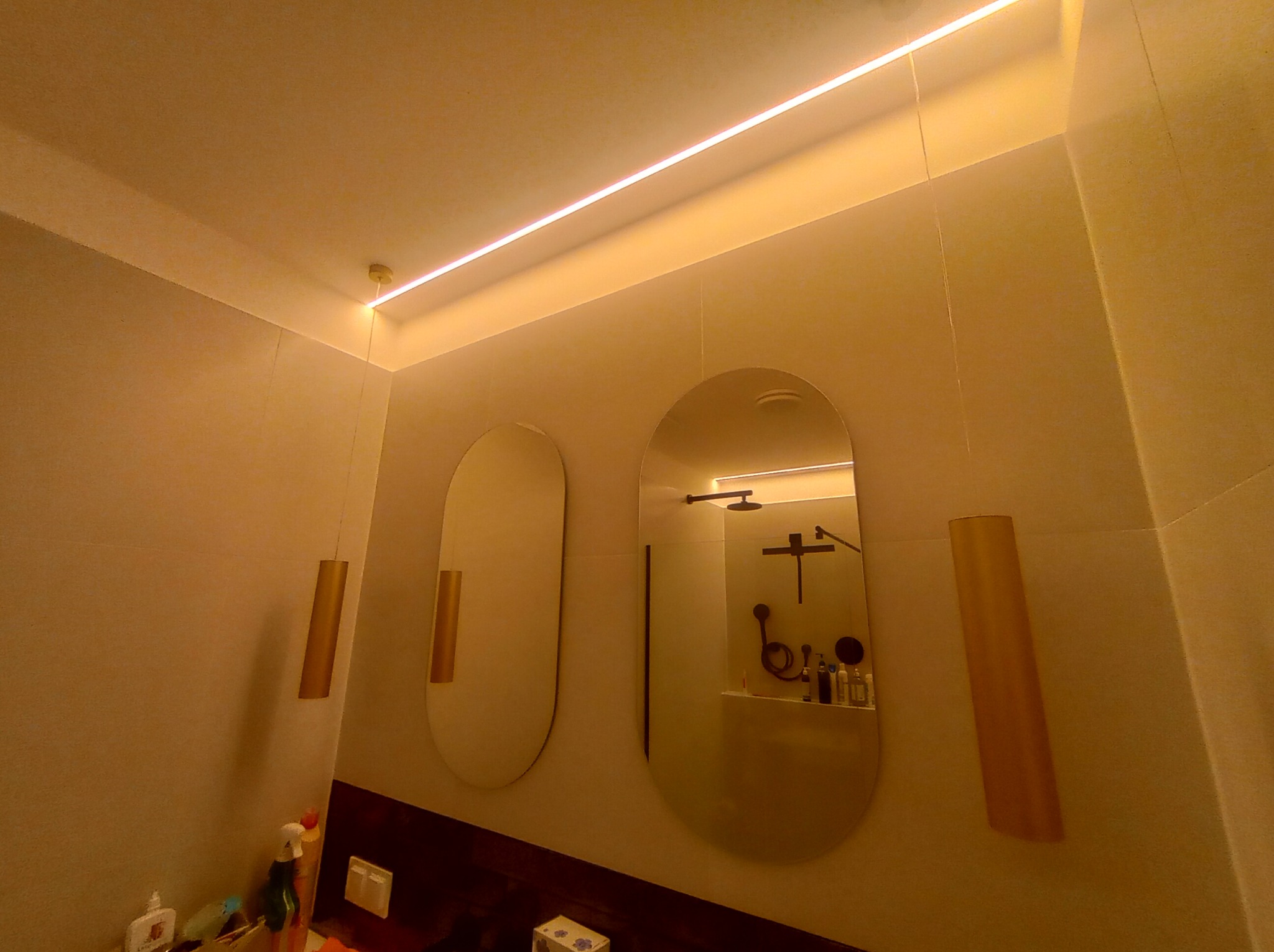 Nowoczesna łazienka z podświetlanym sufitem LED, owalnymi lustrami z dekoracyjnymi lampami wiszącymi i widoczną armaturą prysznicową w odbiciu.