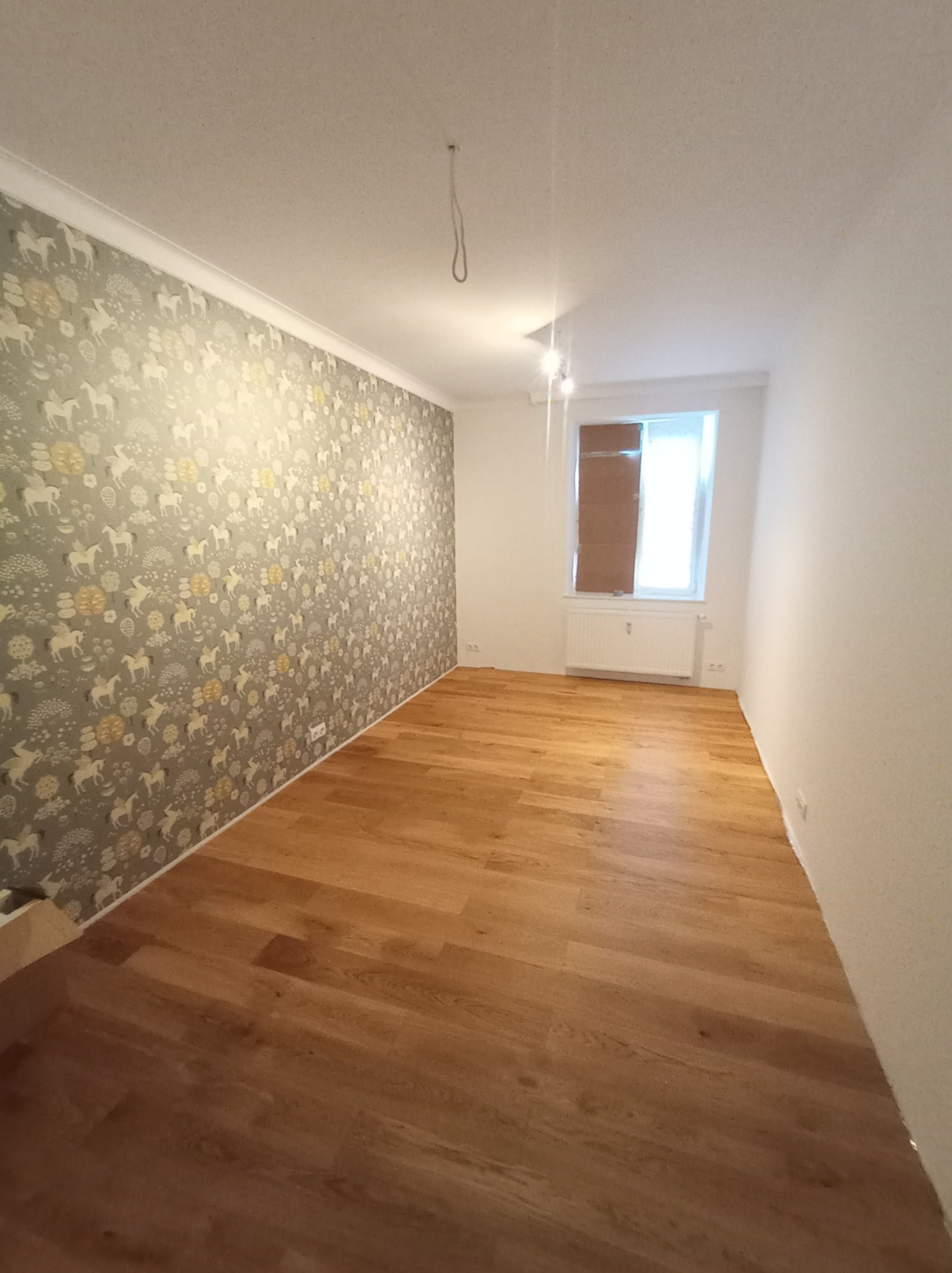 Pokój po remoncie: położony parkiet o ciepłym odcieniu drewna, jedna ściana z tapetą w jasne wzory koni i drzew, pozostałe ściany białe, okno częściowo zasłonięte kartonem.