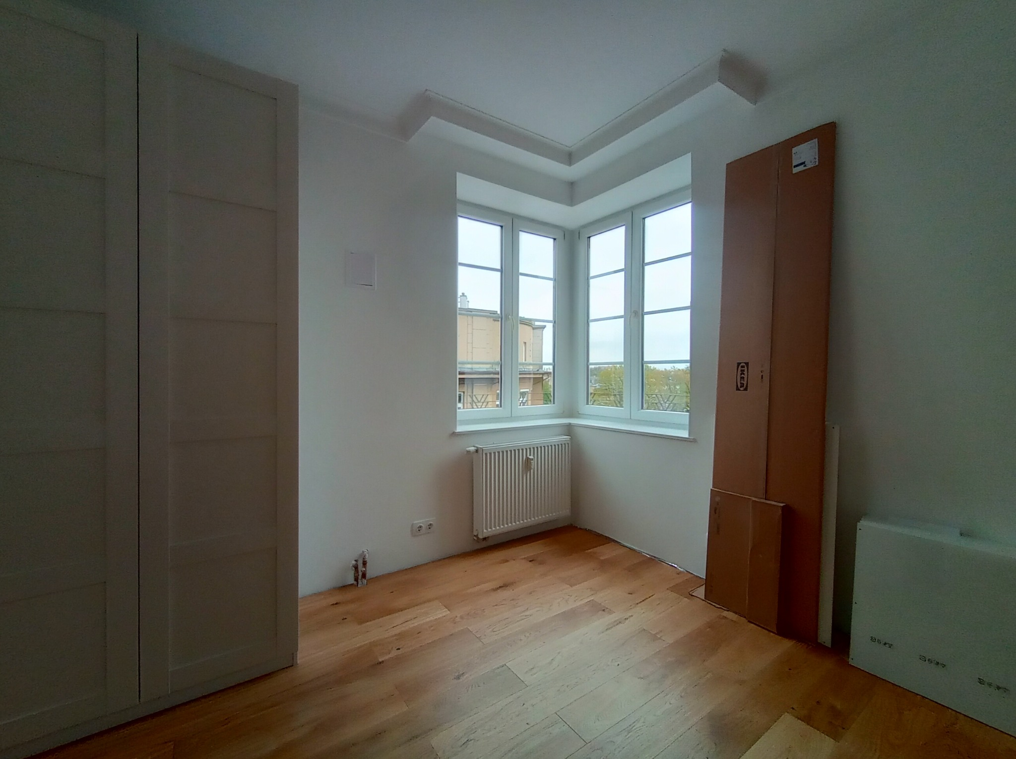 Jasne wnętrze pokoju po remoncie z drewnianą podłogą, białymi ścianami, oknem wykuszowym i grzejnikiem, z elementami do montażu mebli w kartonowych opakowaniach.