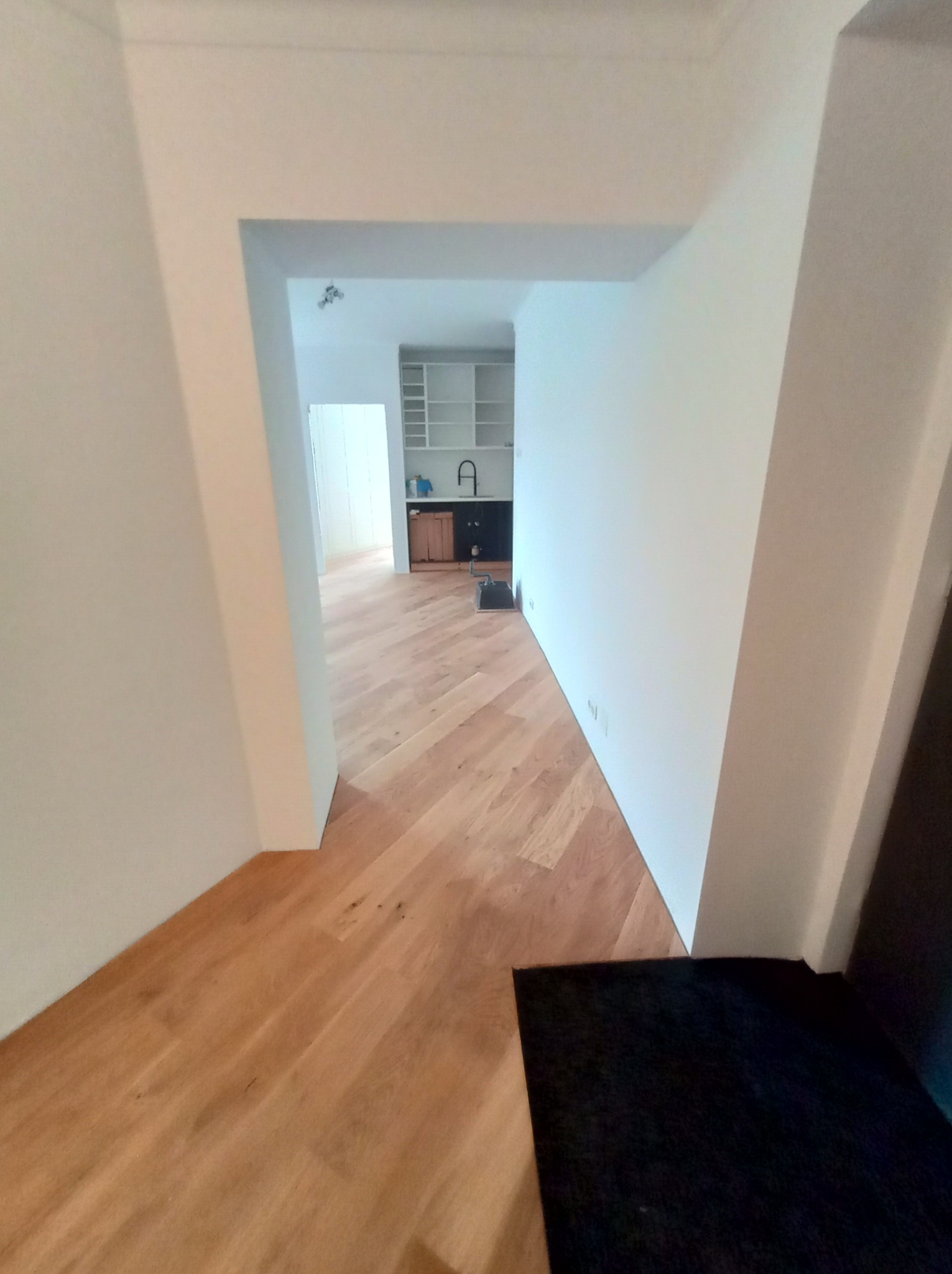 Wykończone wnętrze mieszkania z parkietem ułożonym w jodełkę francuską, widok z korytarza na kuchnię z białymi szafkami i drewnianym blatem. Świeżo pomalowane białe ściany.