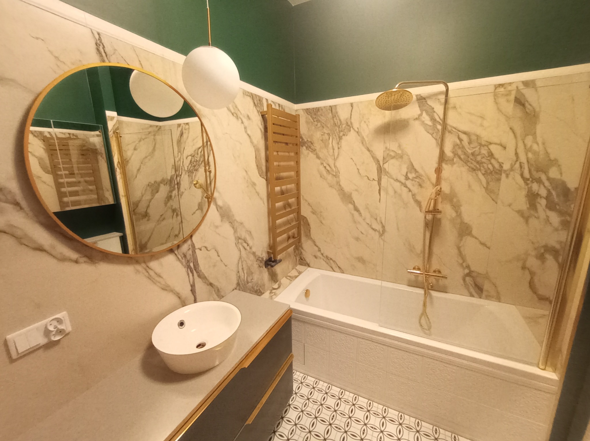 Nowoczesna łazienka z marmurowymi płytkami, okrągłym lustrem w złotej ramie, białą umywalką nablatową, grzejnikiem drabinkowym w kolorze złota i baterią prysznicową w stylu retro.