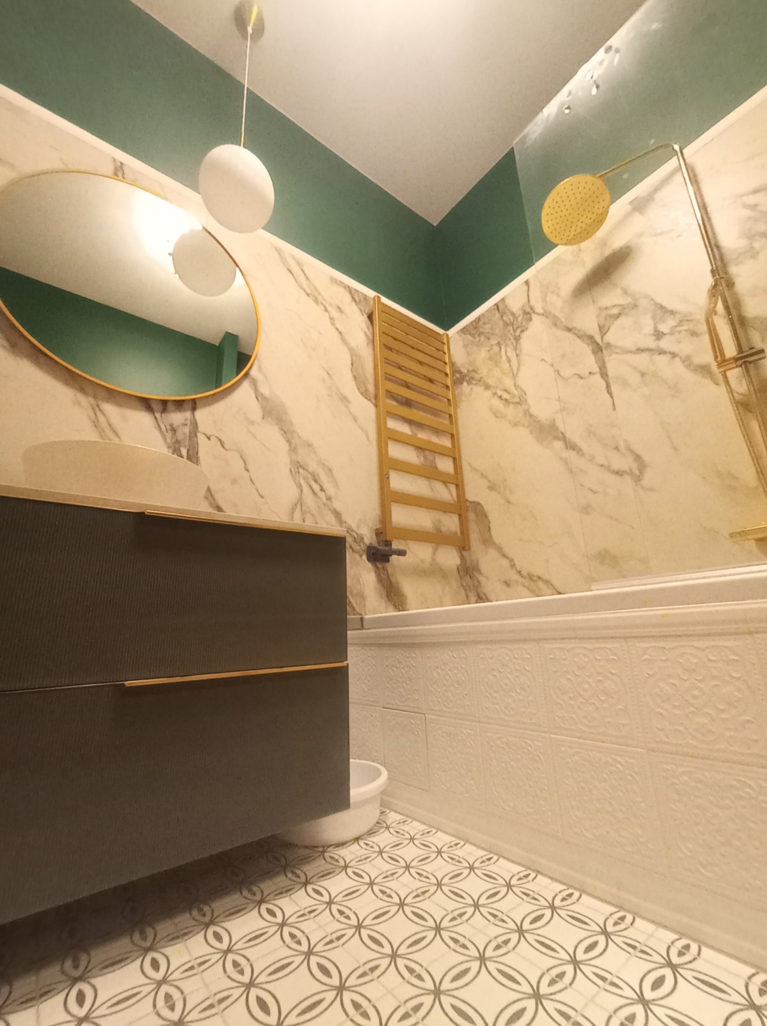 Nowoczesna łazienka z umywalką na szafce w kolorze grafitowym, owalnym lustrem w złotej ramie, grzejnikiem drabinkowym w kolorze złotym i marmurowymi płytkami na ścianach, uzupełniona o białą wannę...