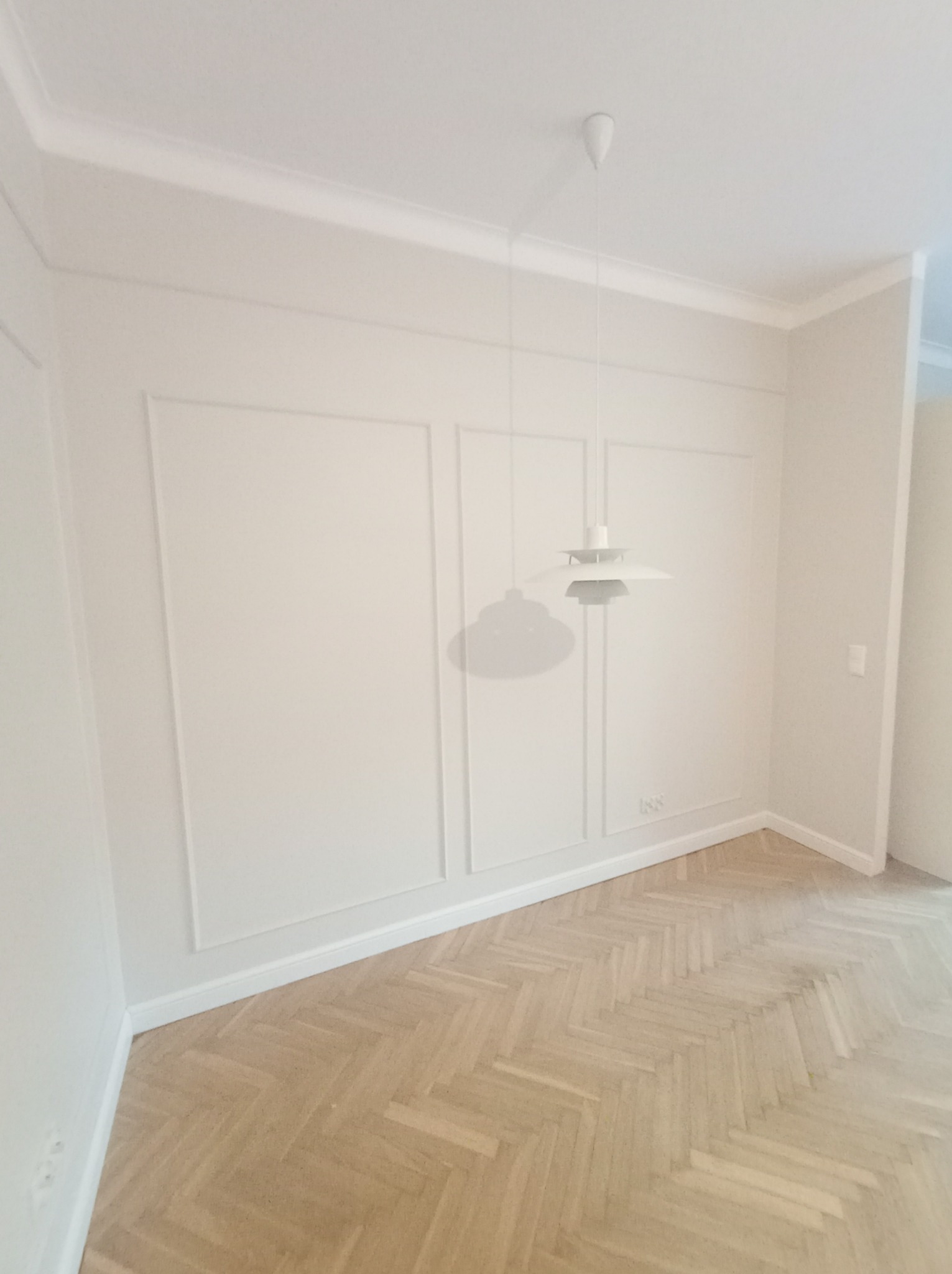 Jasne, minimalistyczne wnętrze po remoncie: parkiet ułożony w jodełkę, białe ściany z subtelnymi listwami dekoracyjnymi, nowoczesna lampa wisząca.