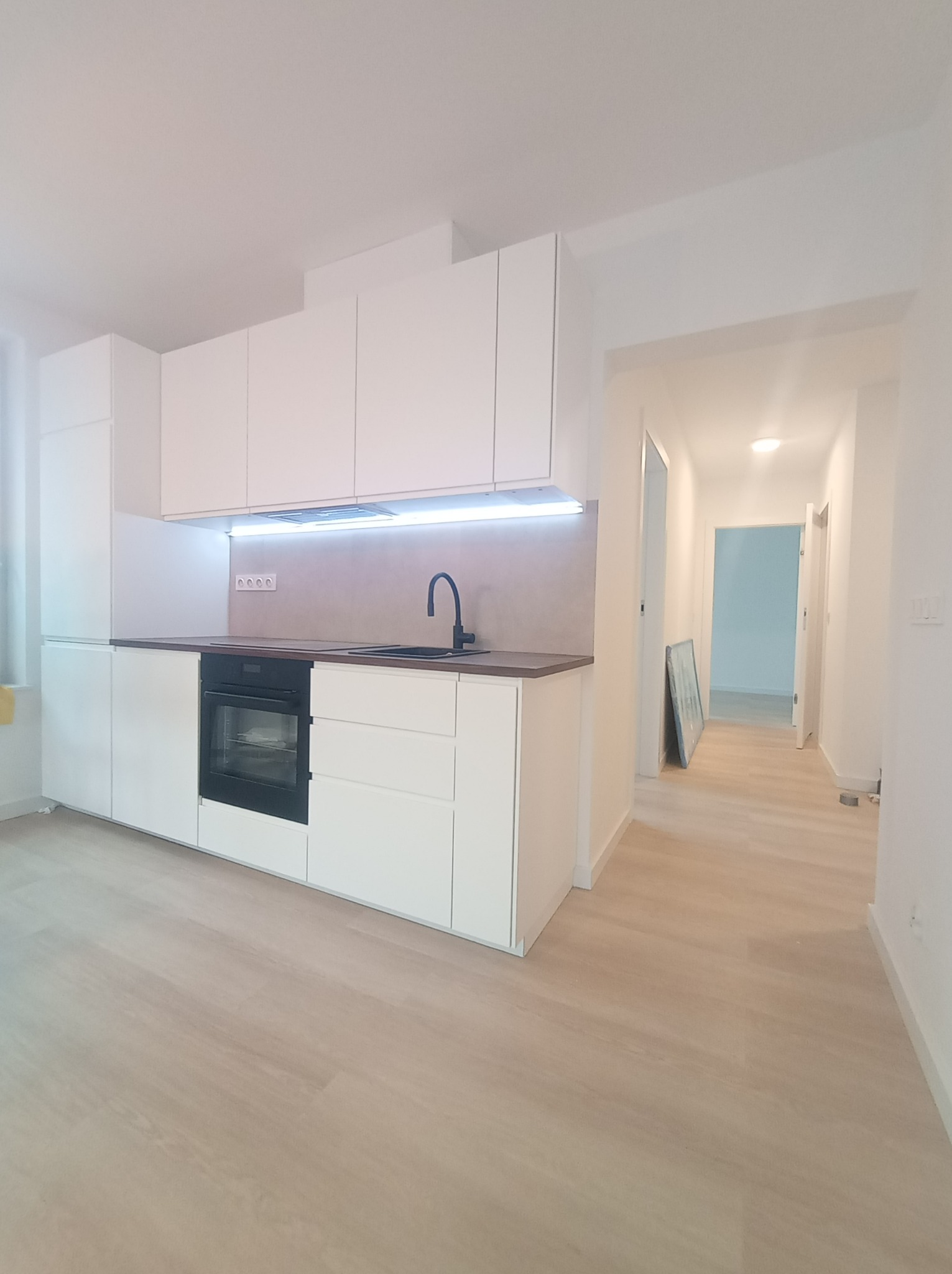 Minimalistyczna, biała zabudowa kuchenna z czarną armaturą i piekarnikiem w nowym mieszkaniu, widok na jasny parkiet i korytarz w tle.