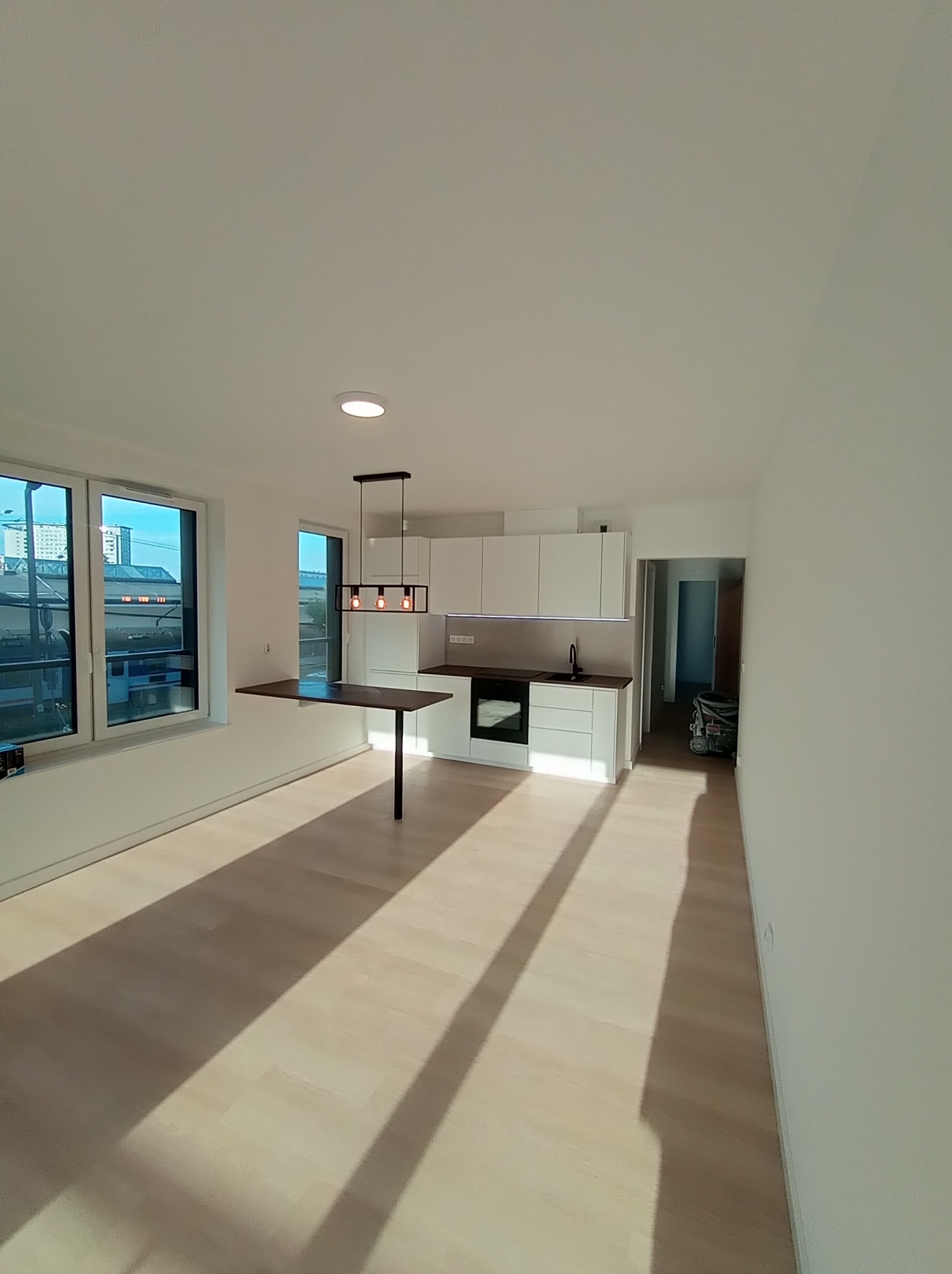 Jasne, przestronne wnętrze mieszkania po remoncie, z białymi ścianami, drewnianą podłogą, nowoczesną kuchnią zintegrowaną z blatem przy oknie, oraz wiszącą lampą z trzema żarówkami.