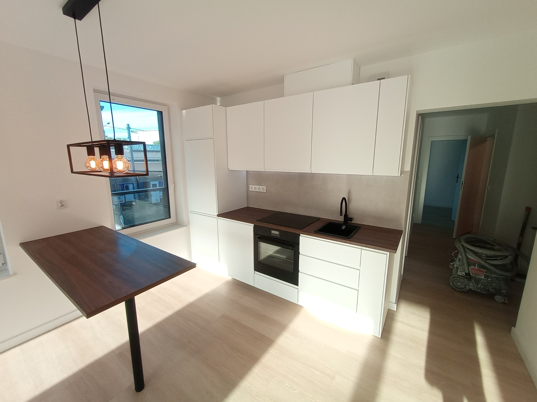 Nowoczesna kuchnia z białymi szafkami, ciemnym blatem i stołem barowym pod industrialną lampą, widoczny odkurzacz przemysłowy w przejściu.