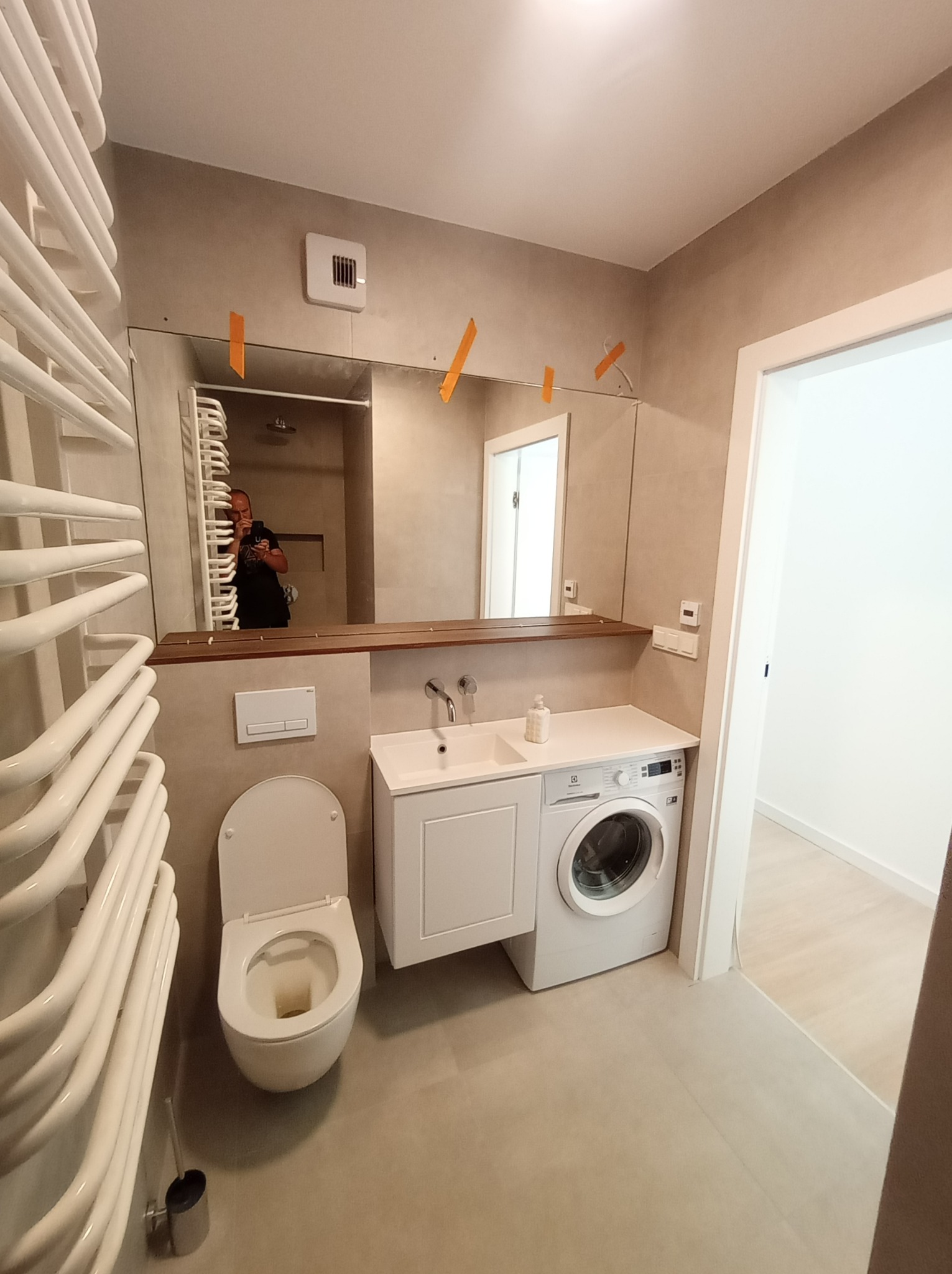 Wykończona łazienka z toaletą, umywalką w zabudowie, pralką oraz grzejnikiem drabinkowym, widoczne taśmy malarskie na lustrze.