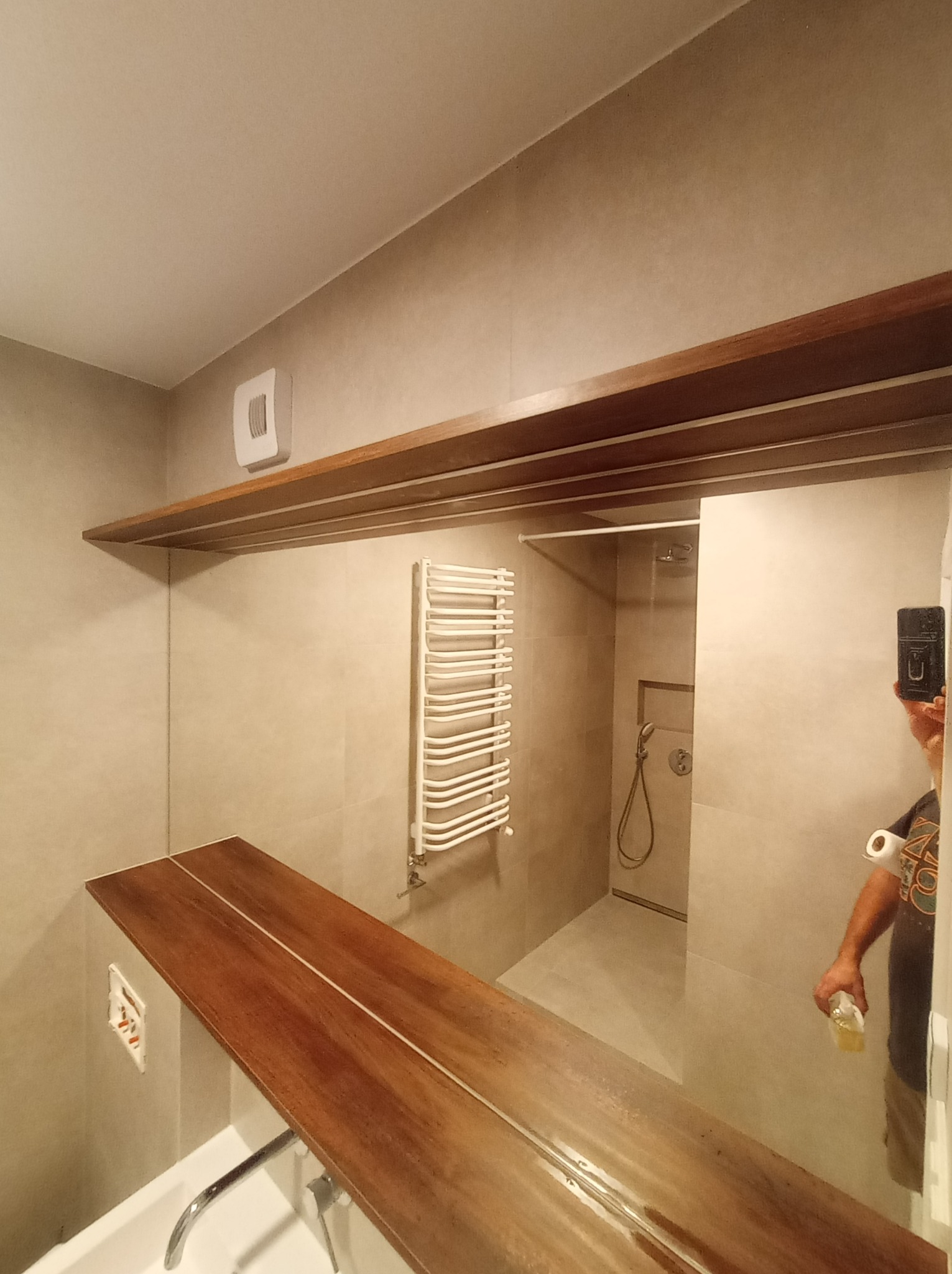 Wykończona łazienka z drewnianym blatem nad umywalką, grzejnikiem drabinkowym i kabiną prysznicową z odpływem liniowym w podłodze, widoczne płytki w odcieniach szarości oraz drewniana półka...