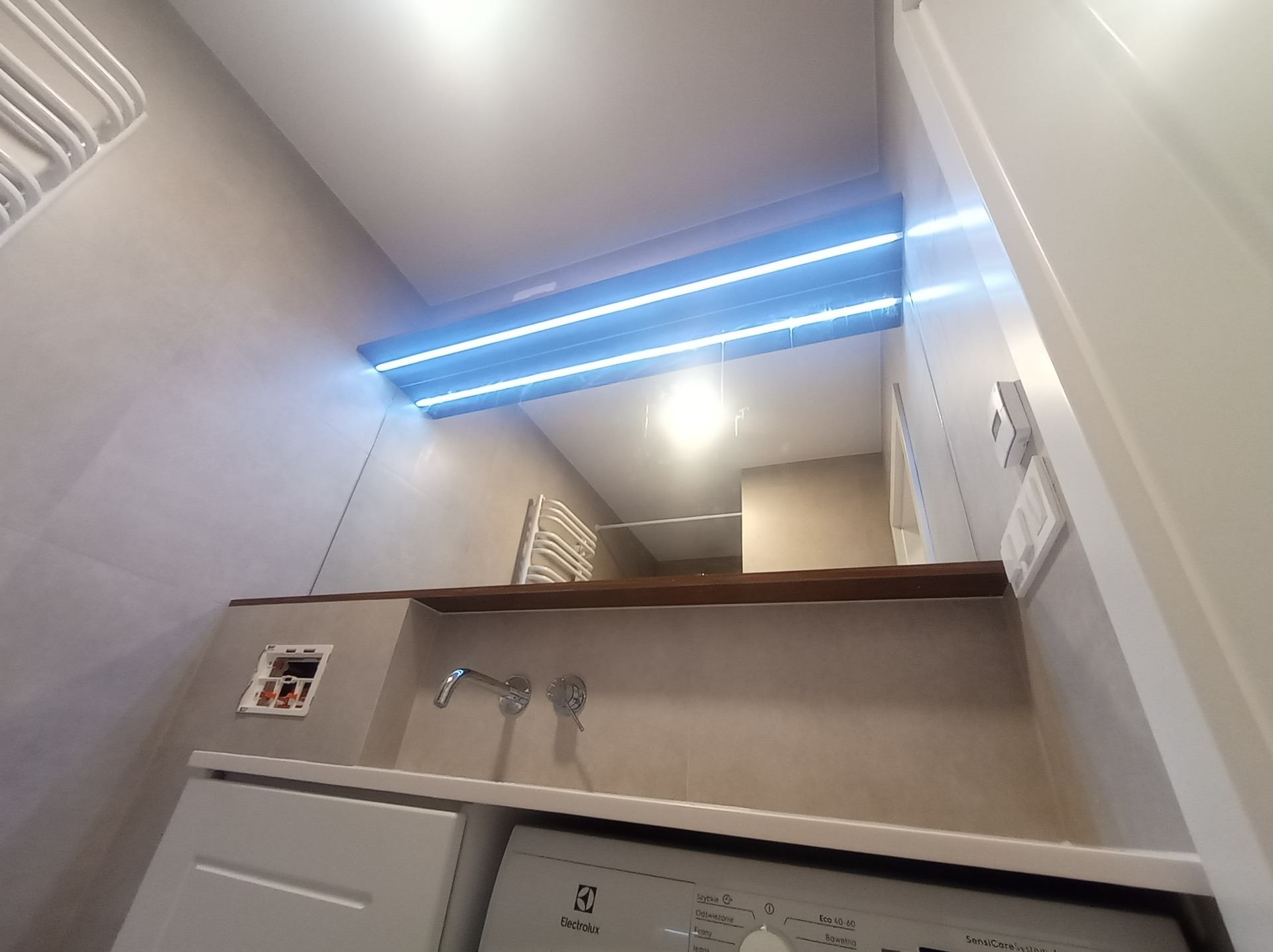 Ukończona łazienka z podświetlanym lustrem, baterią umywalkową, pralką Electrolux i niezamaskowanym gniazdkiem elektrycznym.