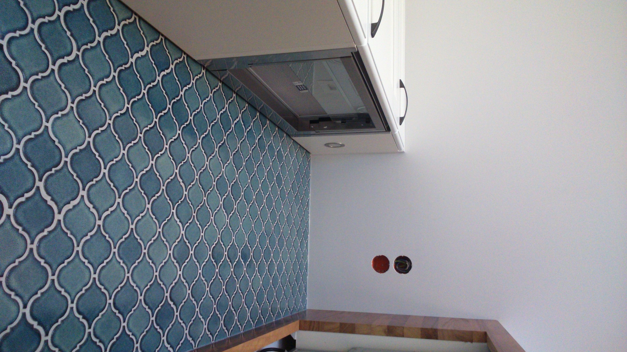 Ściana wyłożona mozaiką w odcieniach turkusu i bieli, z widocznym okapem kuchennym i fragmentem blatu drewnianego, na ścianie gniazdka elektryczne bez osprzętu.