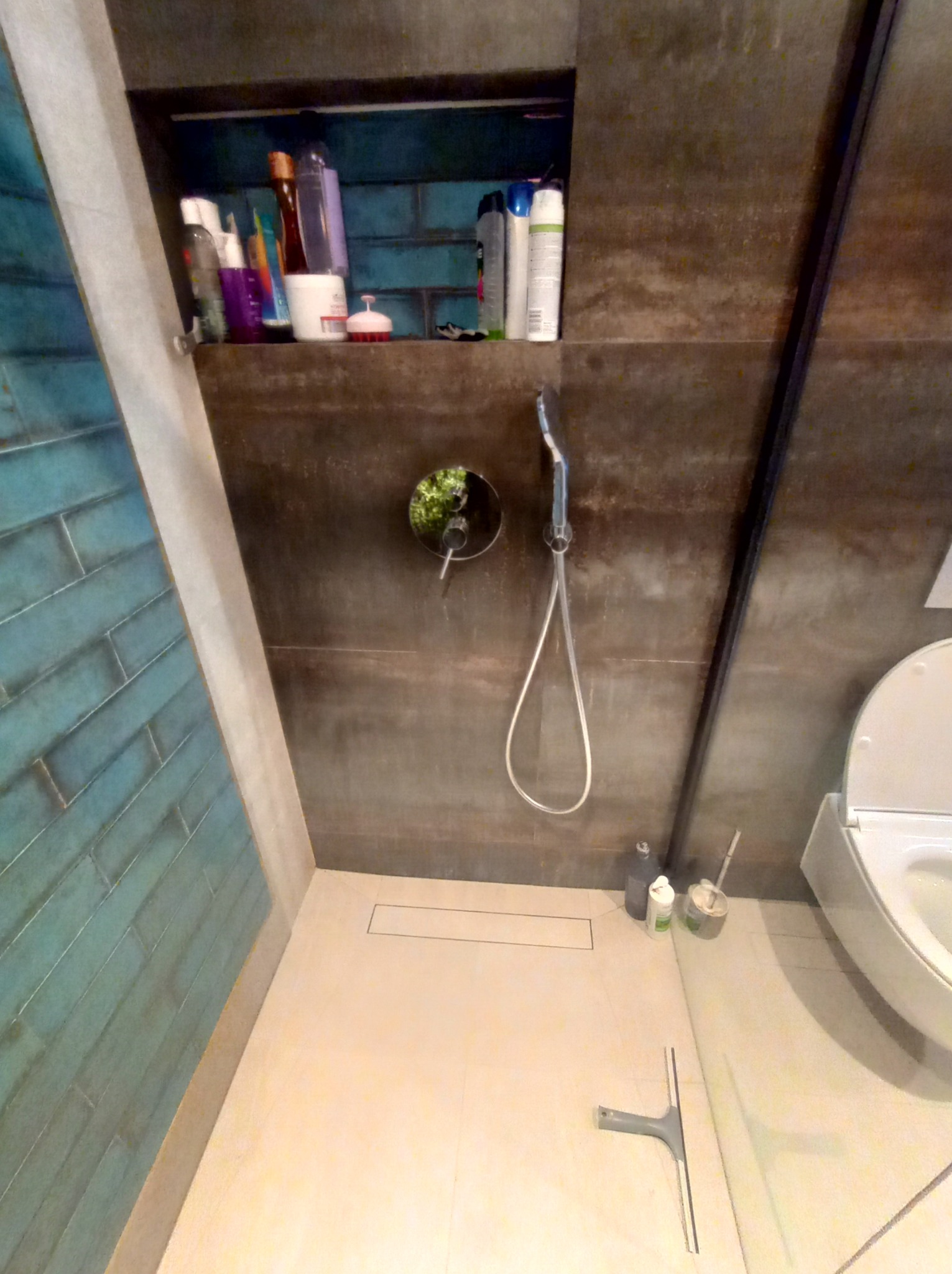 Nowoczesna łazienka z turkusową ścianą z płytek, ciemną ścianą z ukrytym prysznicem i półką z kosmetykami, oraz odpływem liniowym w podłodze. Widoczna toaleta i akcesoria do sprzątania.