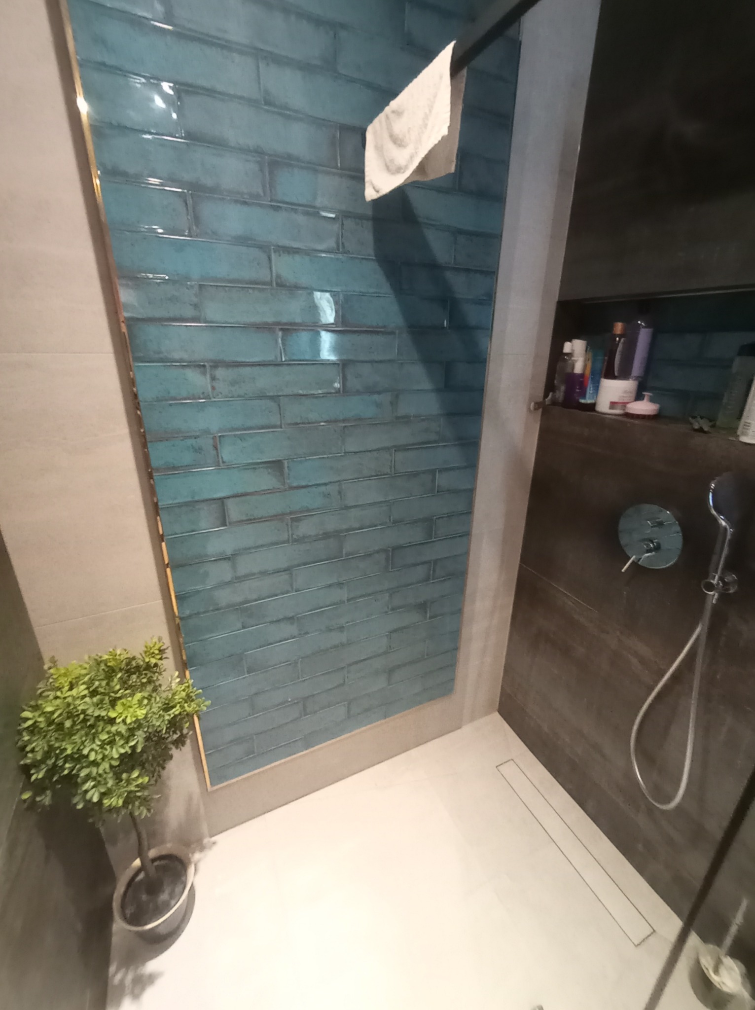 Nowoczesna kabina prysznicowa z turkusowymi, błyszczącymi płytkami ułożonymi w cegiełkę, szara ściana, biała posadzka z odpływem liniowym, roślina doniczkowa w rogu, półka z kosmetykami i słuchawka...